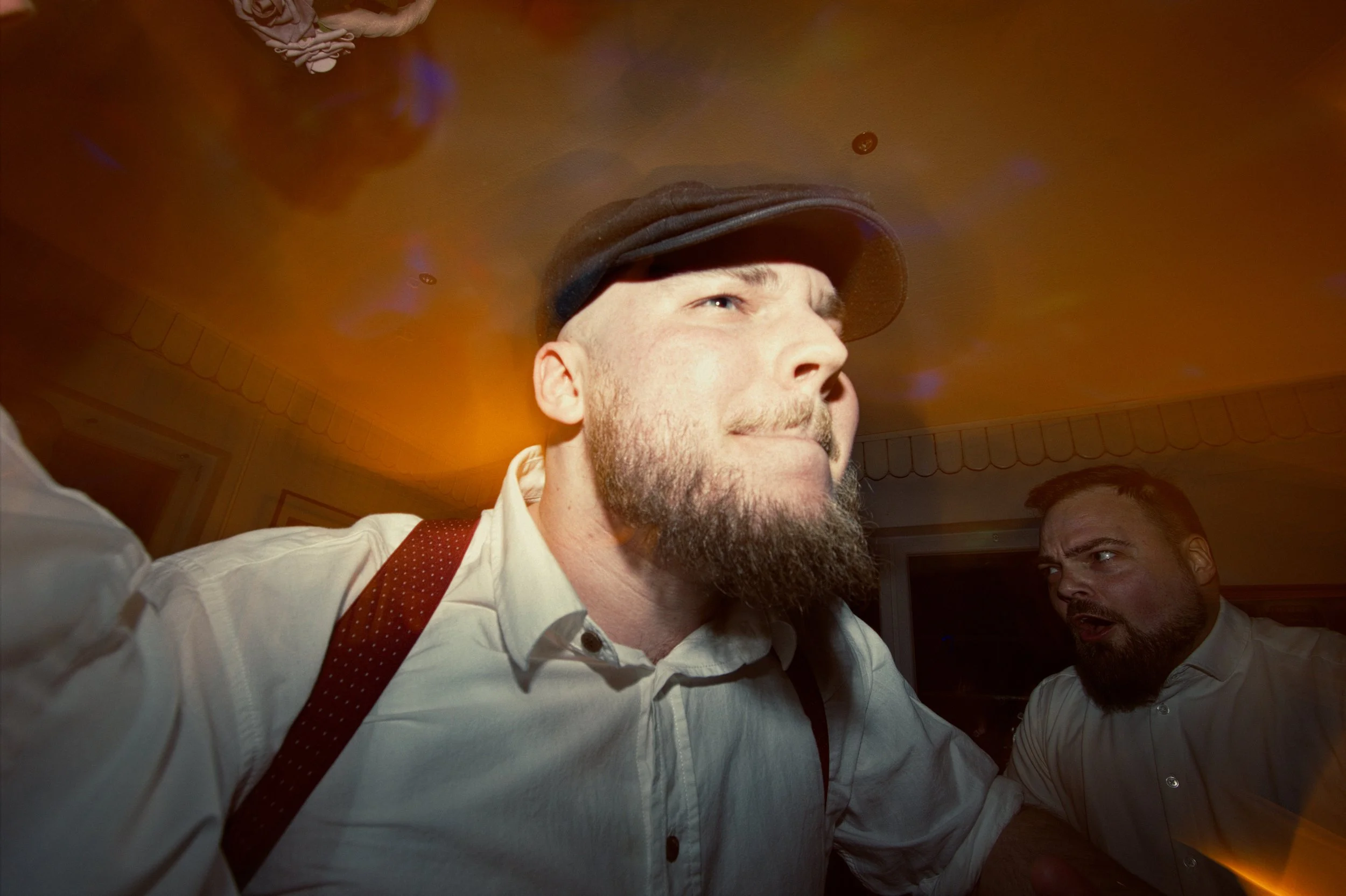 A man with a beard and a cap making a serious face, with another man behind him looking surprised or angry, in an indoor setting with warm lighting.