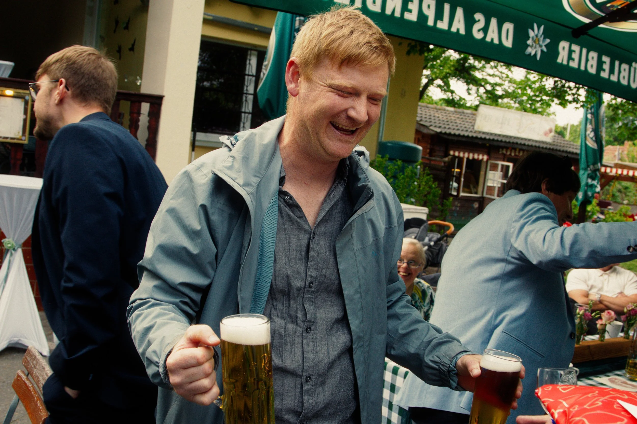 A man with red hair and a gray jacket holding two glasses of beer, smiling at an outdoor gathering.