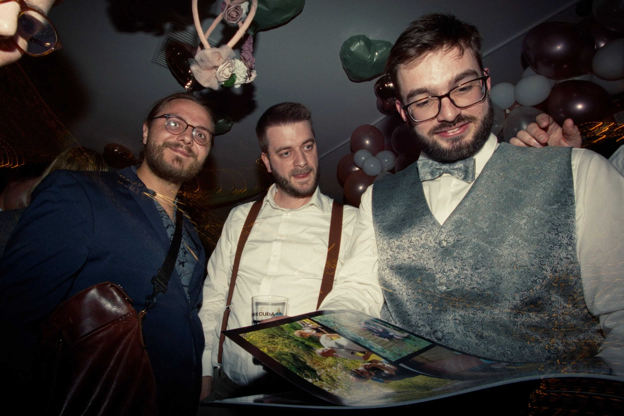 Three men at a celebration or party, looking at a photo album or book, with balloons and decorations in the background.