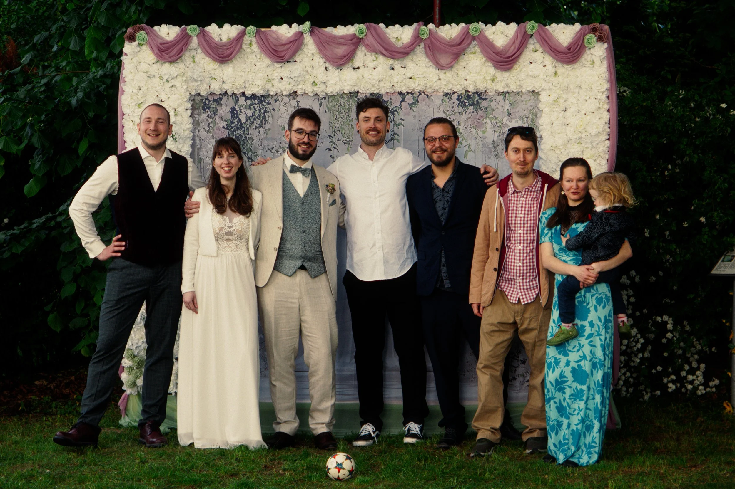 Group of eight people standing outdoors in front of a floral backdrop, smiling, for a celebration or gathering.