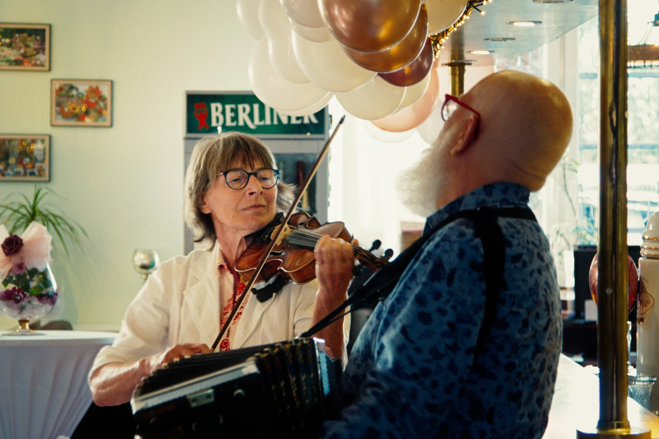 An elderly woman playing the violin and an elderly man with glasses playing an accordion in a decorated room with balloons and artwork.