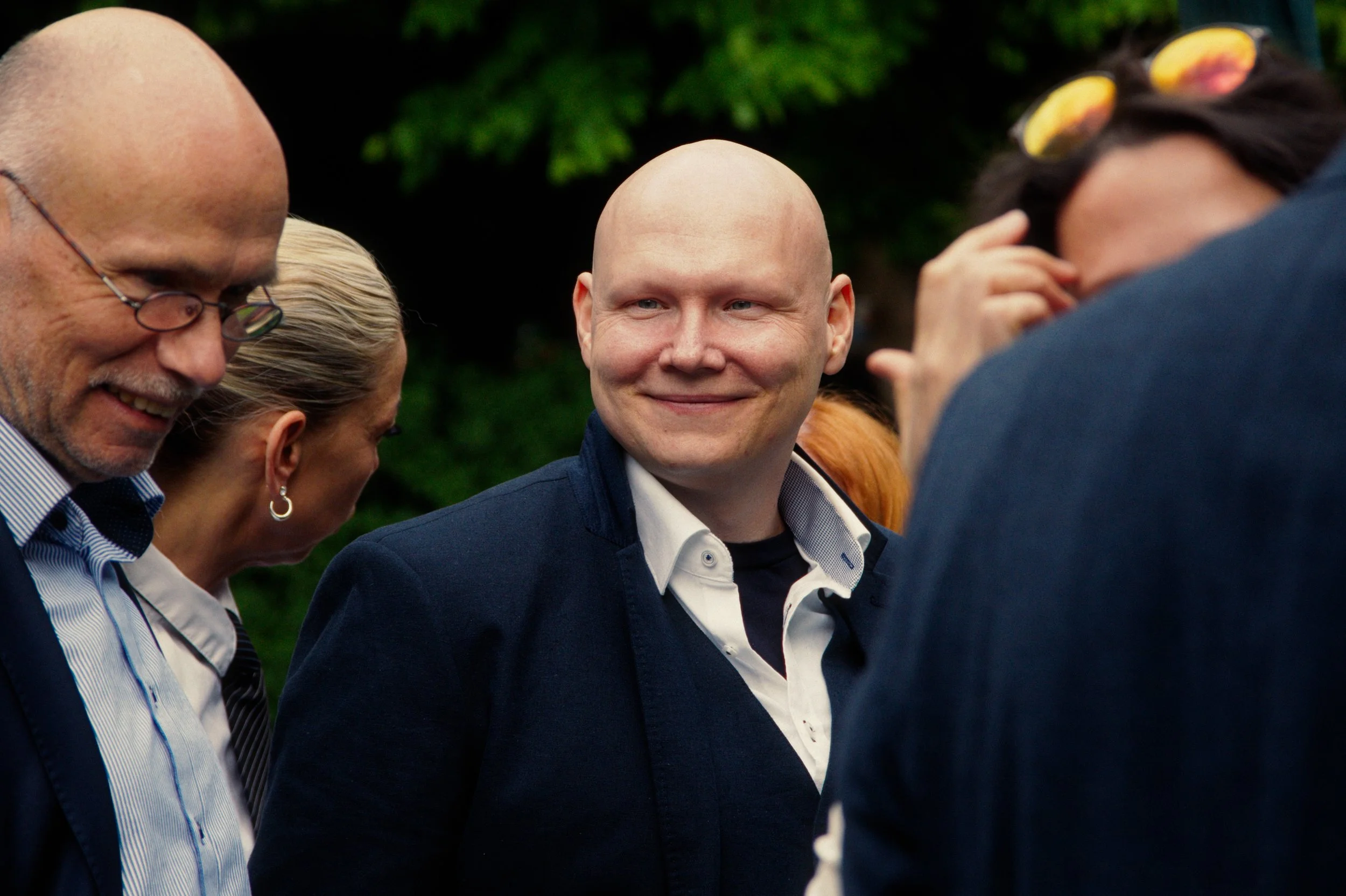 A group of people outdoors, with a man in the center smiling and engaging in conversation. The background is green, indicating a park or garden setting.