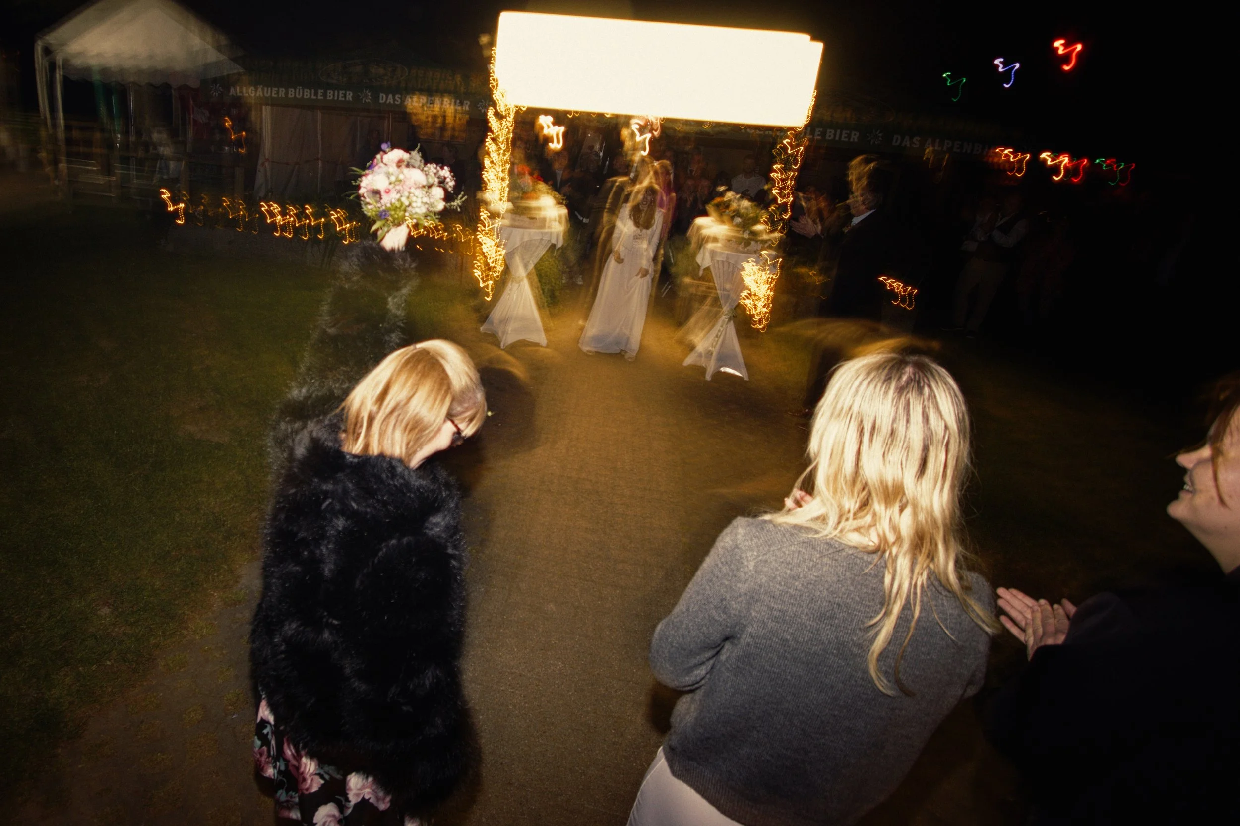 Three women standing outside at night, with wedding reception tables and a bride in a white dress in the background, illuminated by string lights and decorative lighting.