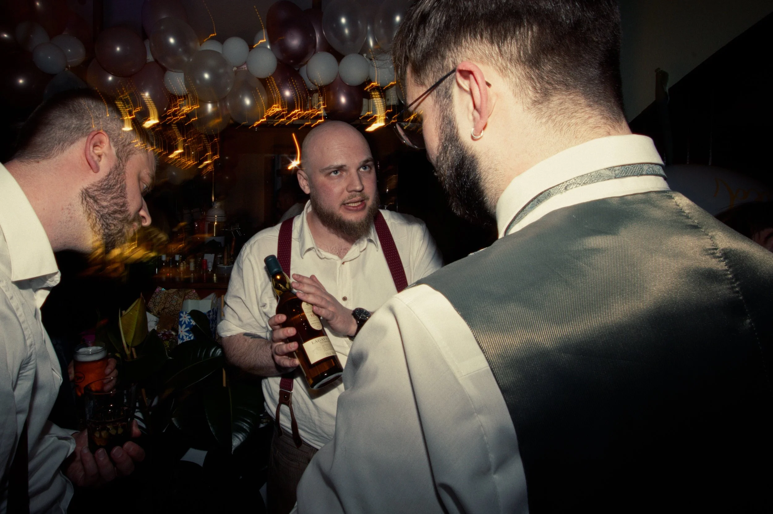 Three men engaged in conversation at a party, one holding a bottle of alcohol, with balloons and festive lighting in the background.