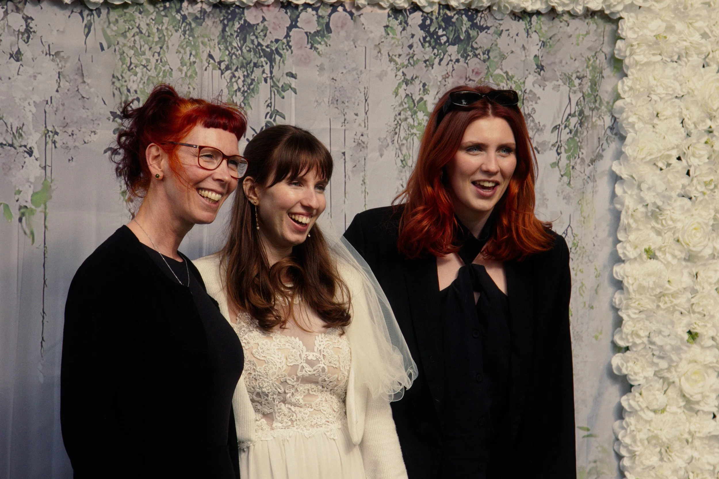 Three women at a wedding, smiling and posing for a photo in front of a floral backdrop. The woman in the middle is wearing a wedding dress, and the others are in black and dark clothing.