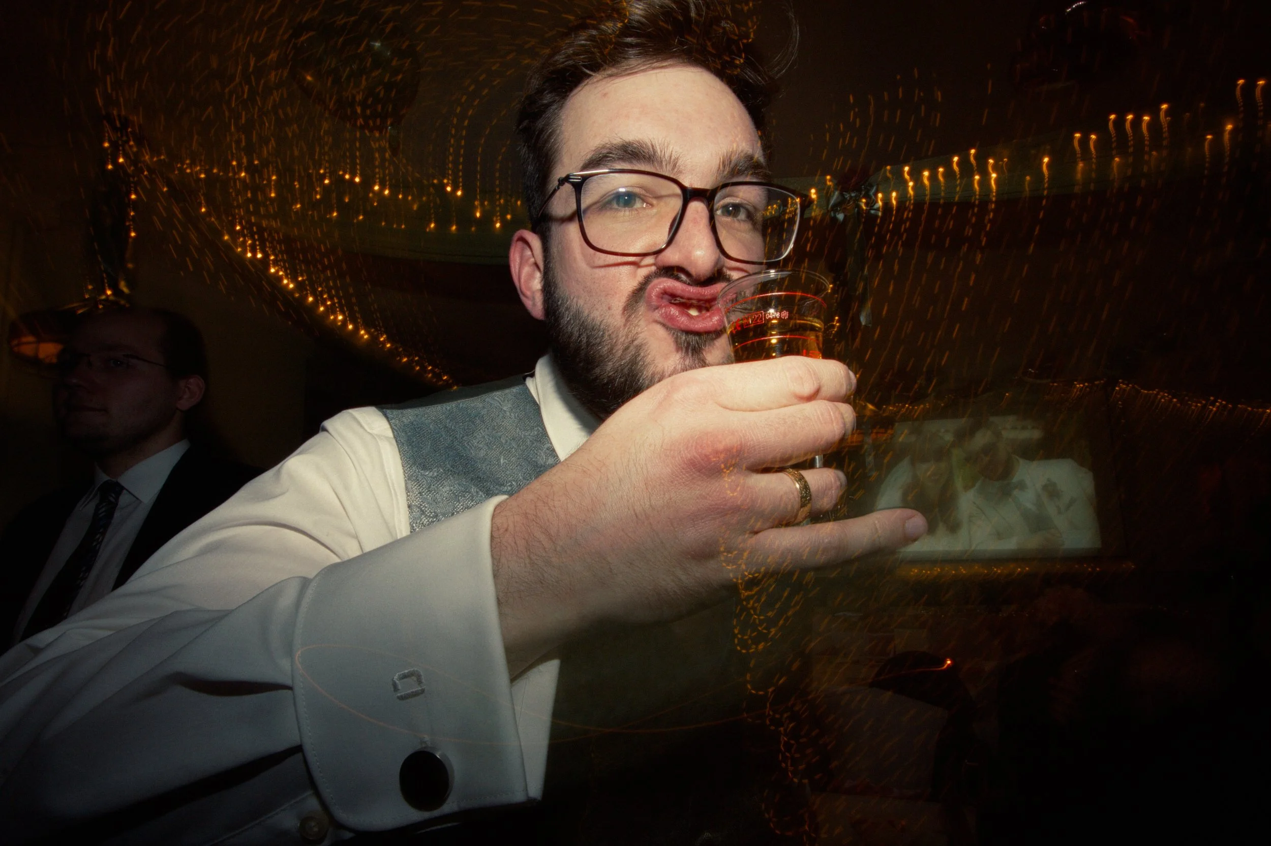 Man with glasses and beard holding a glass of drink at a party, with blurry lights in the background.