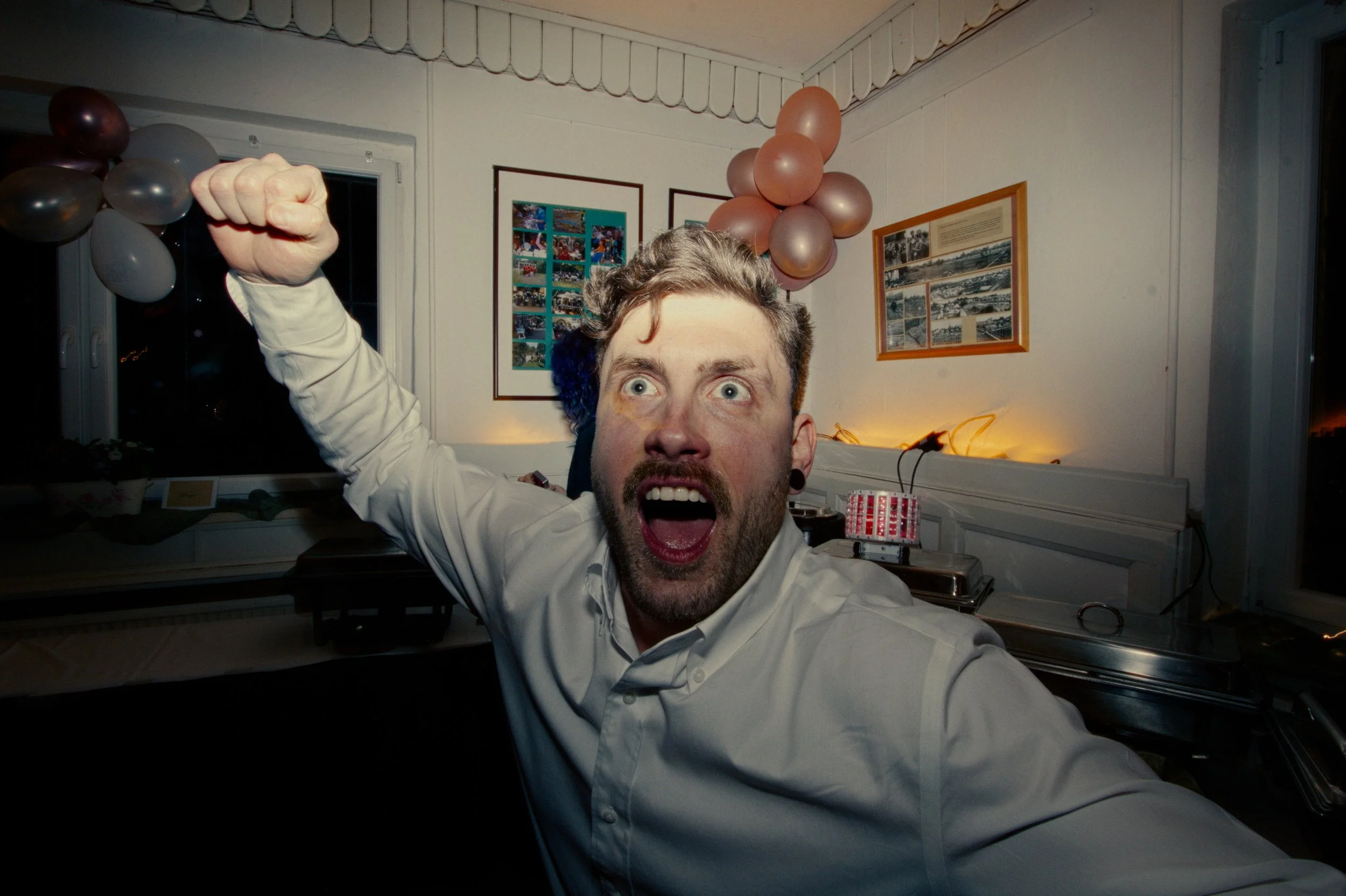 A man with an excited expression at a celebration, raising his fist, with balloons and framed photos on the wall behind him.