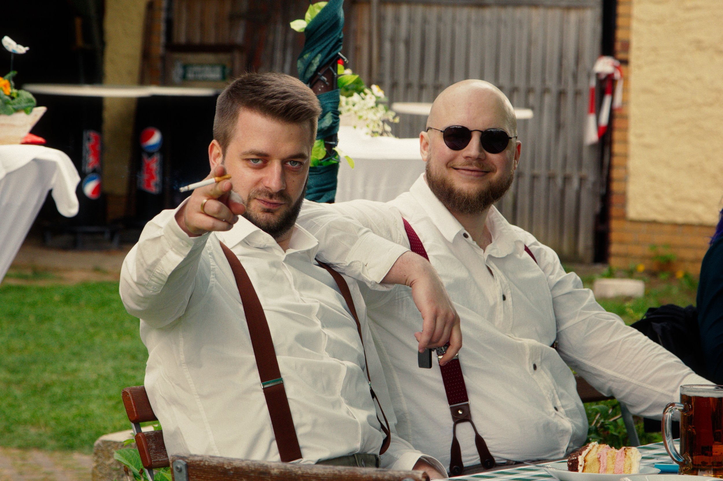 Two men sitting at a table outdoors, one pointing at the camera and the other smiling with sunglasses, both dressed in white shirts and suspenders, with a slice of cake and a drink on the table.
