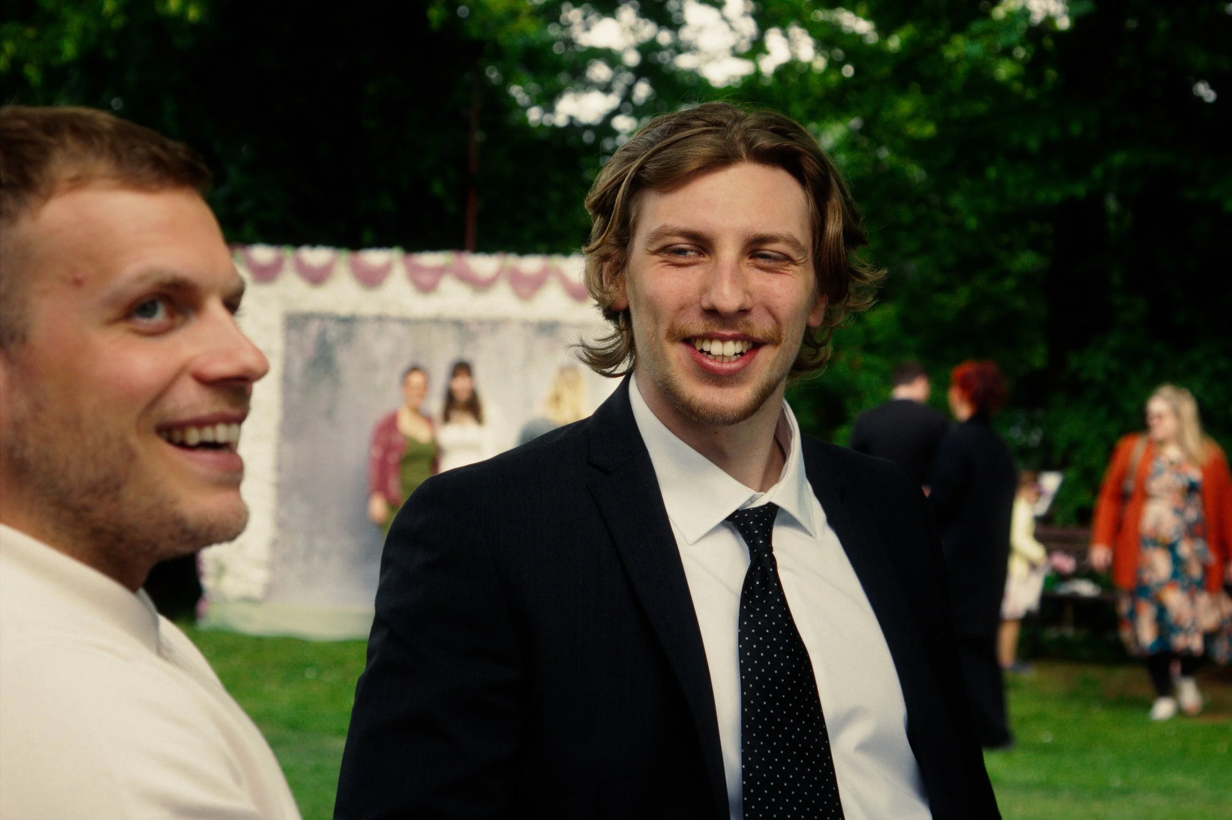 Two men smiling and talking outdoors, with a group of people and a decorated backdrop in the background, likely at a celebration or party.