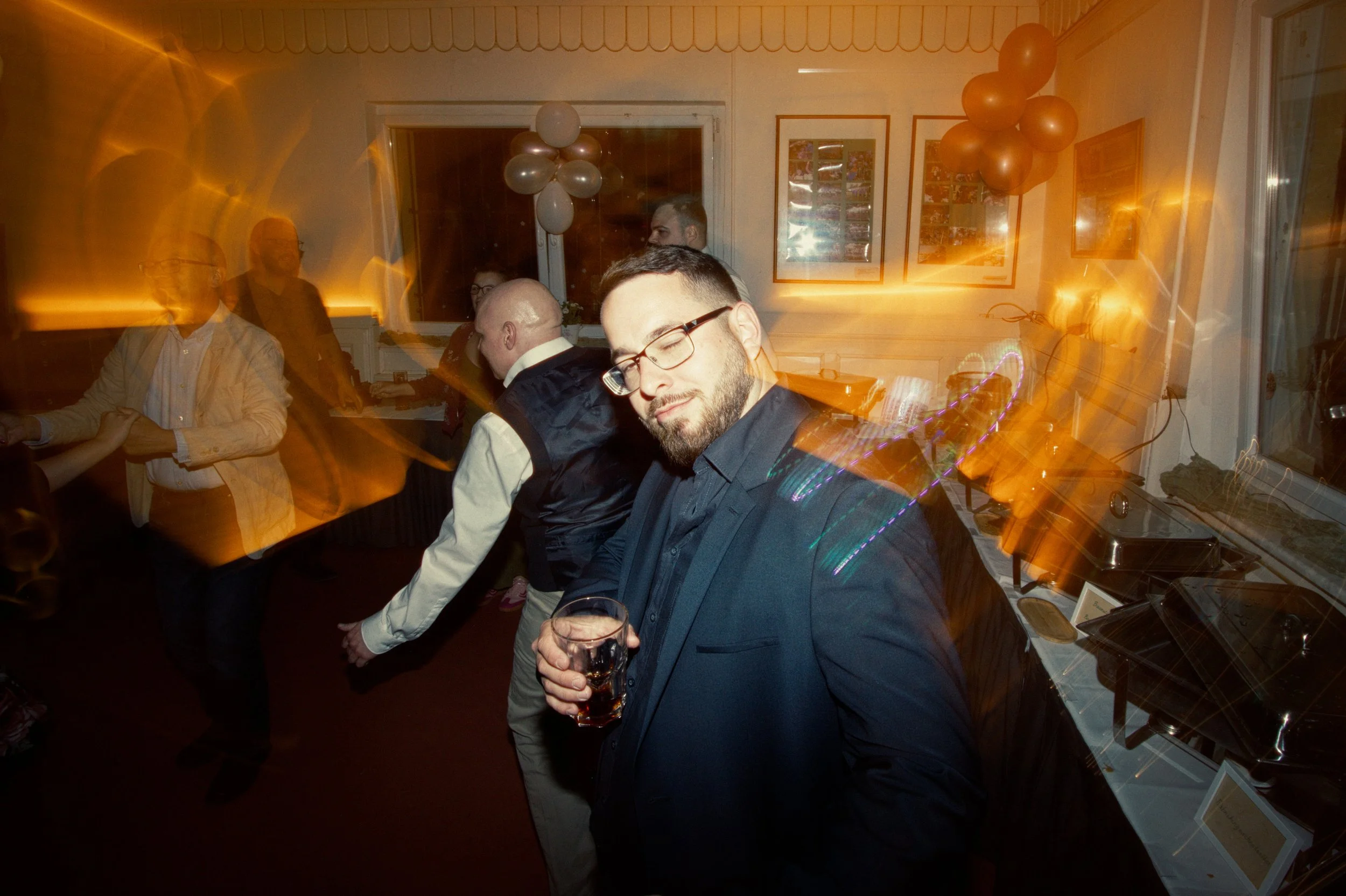 A man with glasses and a beard in a dark suit holding a drink, at a party with blurred dancing figures, balloons, and party decorations.