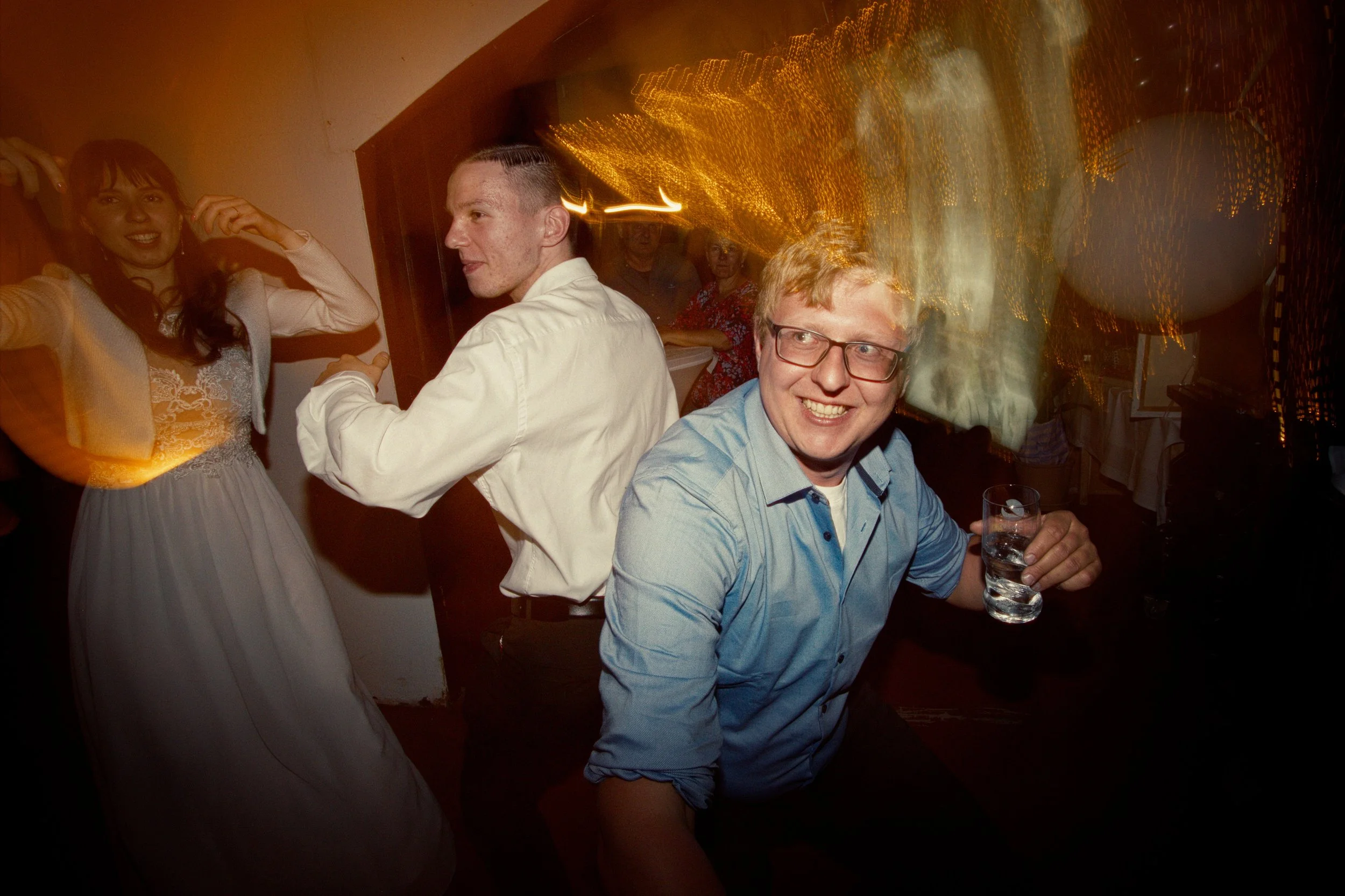 People dancing and having fun at a party, with a man holding a glass of drink smiling at the camera and others dancing in the background.