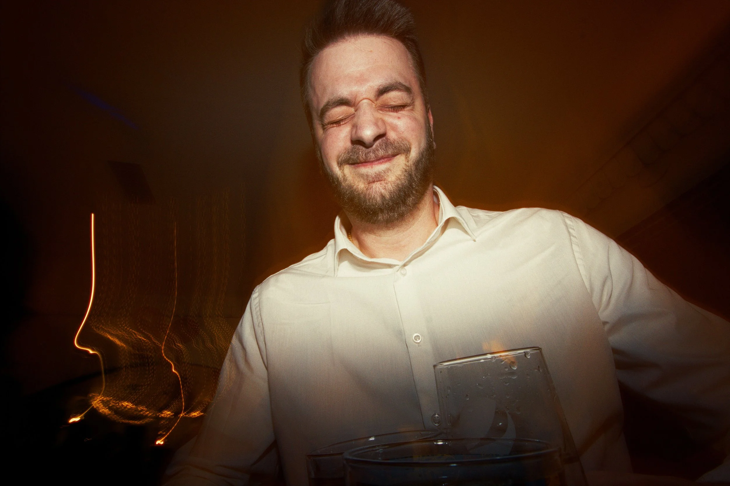 A smiling man with closed eyes and a beard holding a glass of water at an indoor event with warm lighting.