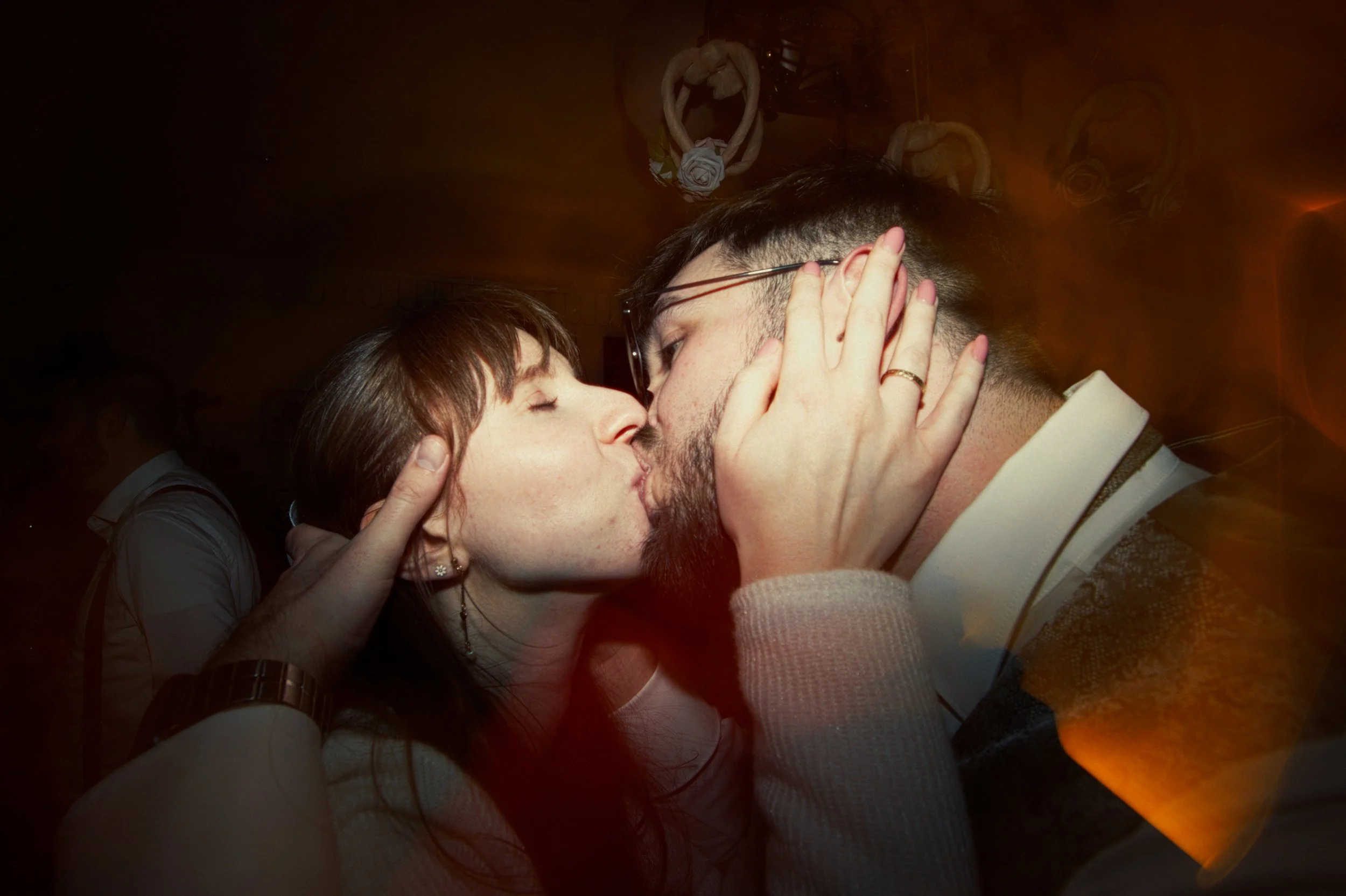 A couple sharing a kiss at a social gathering, holding each other's faces with hands around their cheeks, with blurred background.