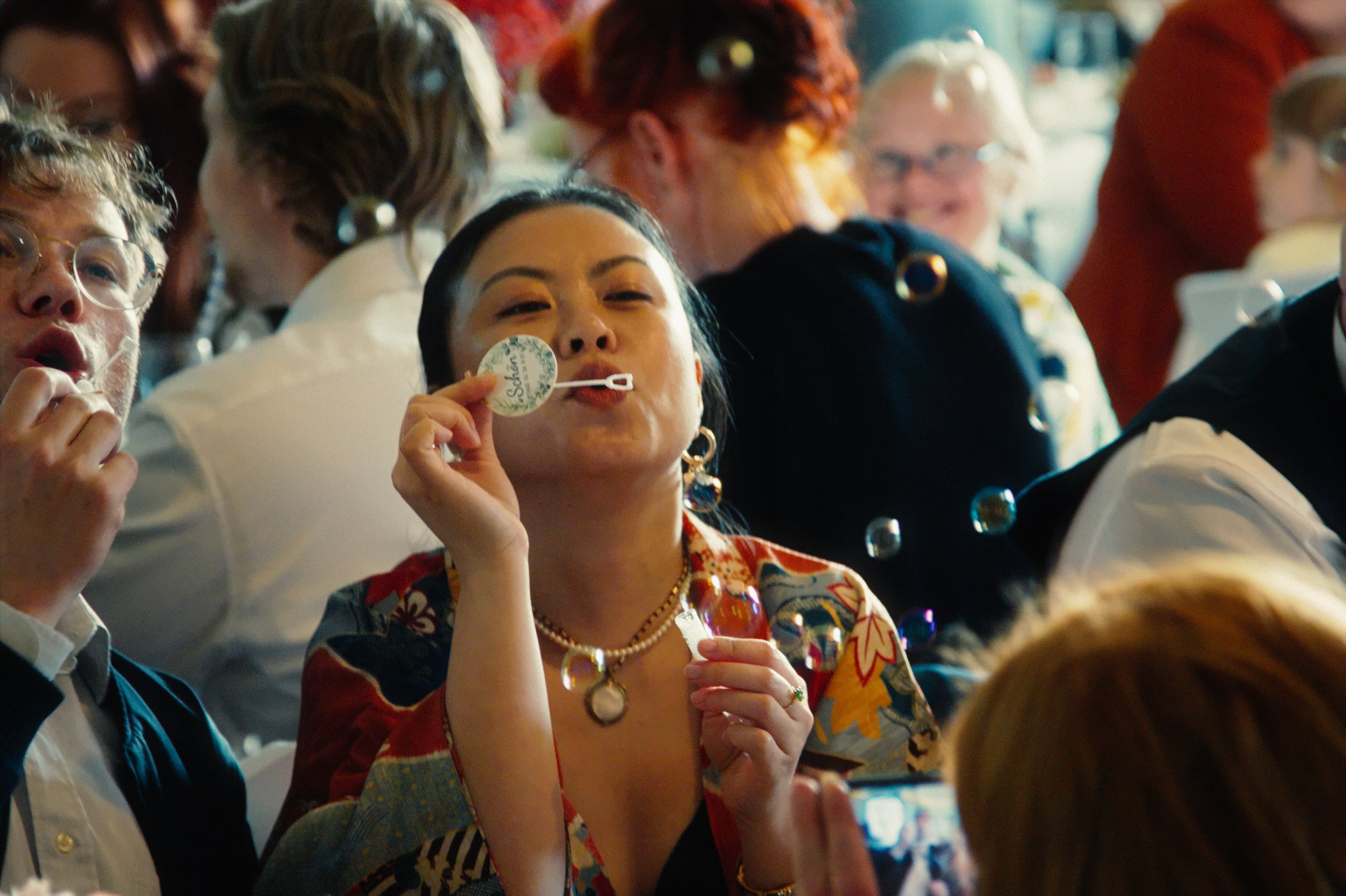 A woman at a social gathering blowing bubbles, holding a bubble wand near her lips, with multiple bubbles floating in the air around her, surrounded by other people in casual and festive attire.