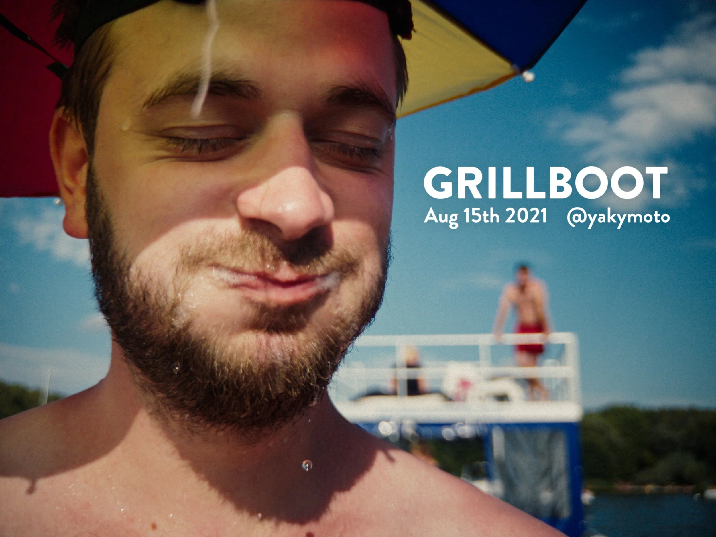 Close-up of a man with a beard and closed eyes smiling, wearing a multicolored umbrella hat, with a boat and a person standing on the boat in the background under a partly cloudy sky.