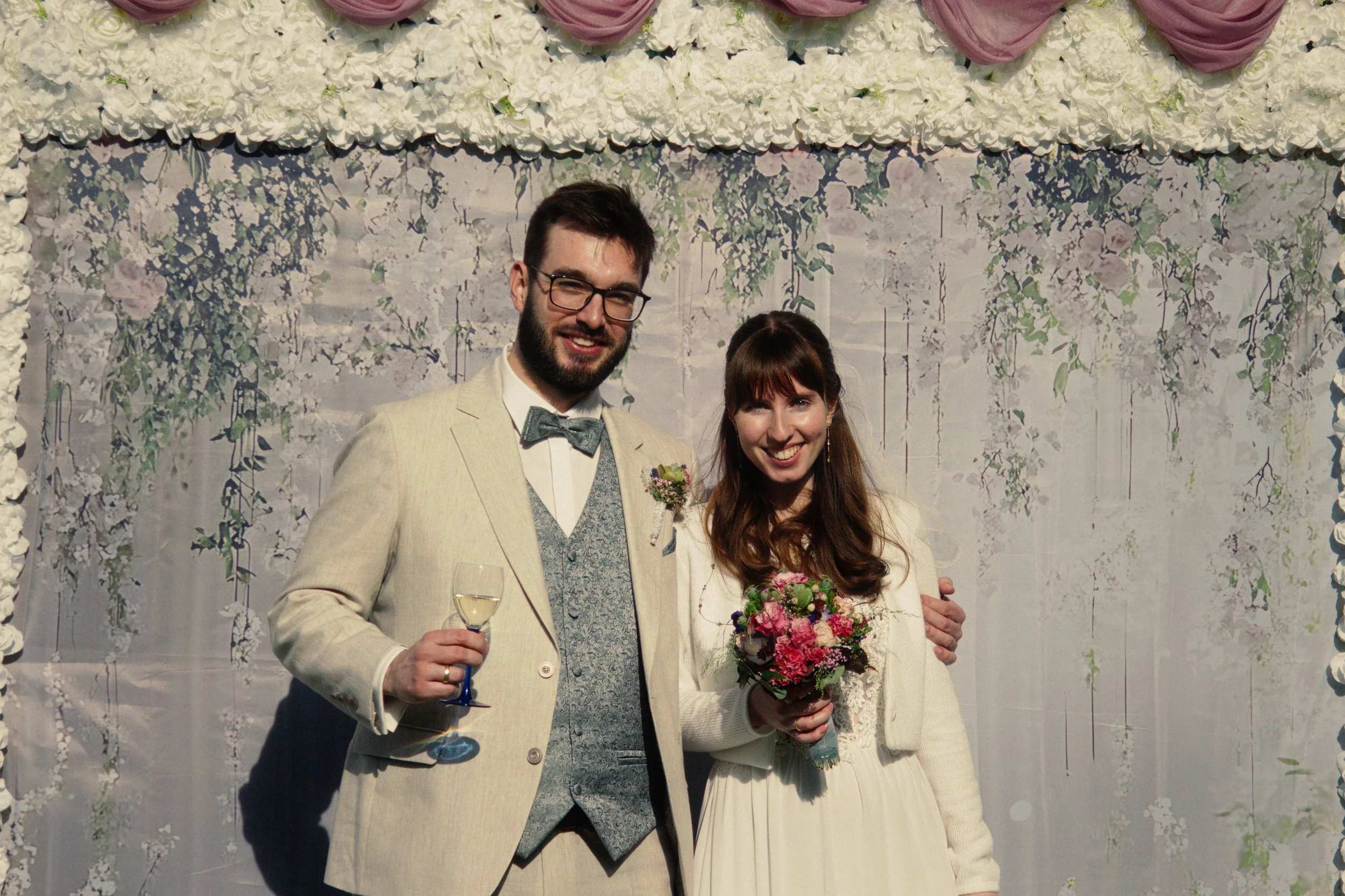 A man and woman at their wedding, smiling, with the man holding a glass of champagne and the woman holding a bouquet of flowers, standing in front of a floral backdrop.