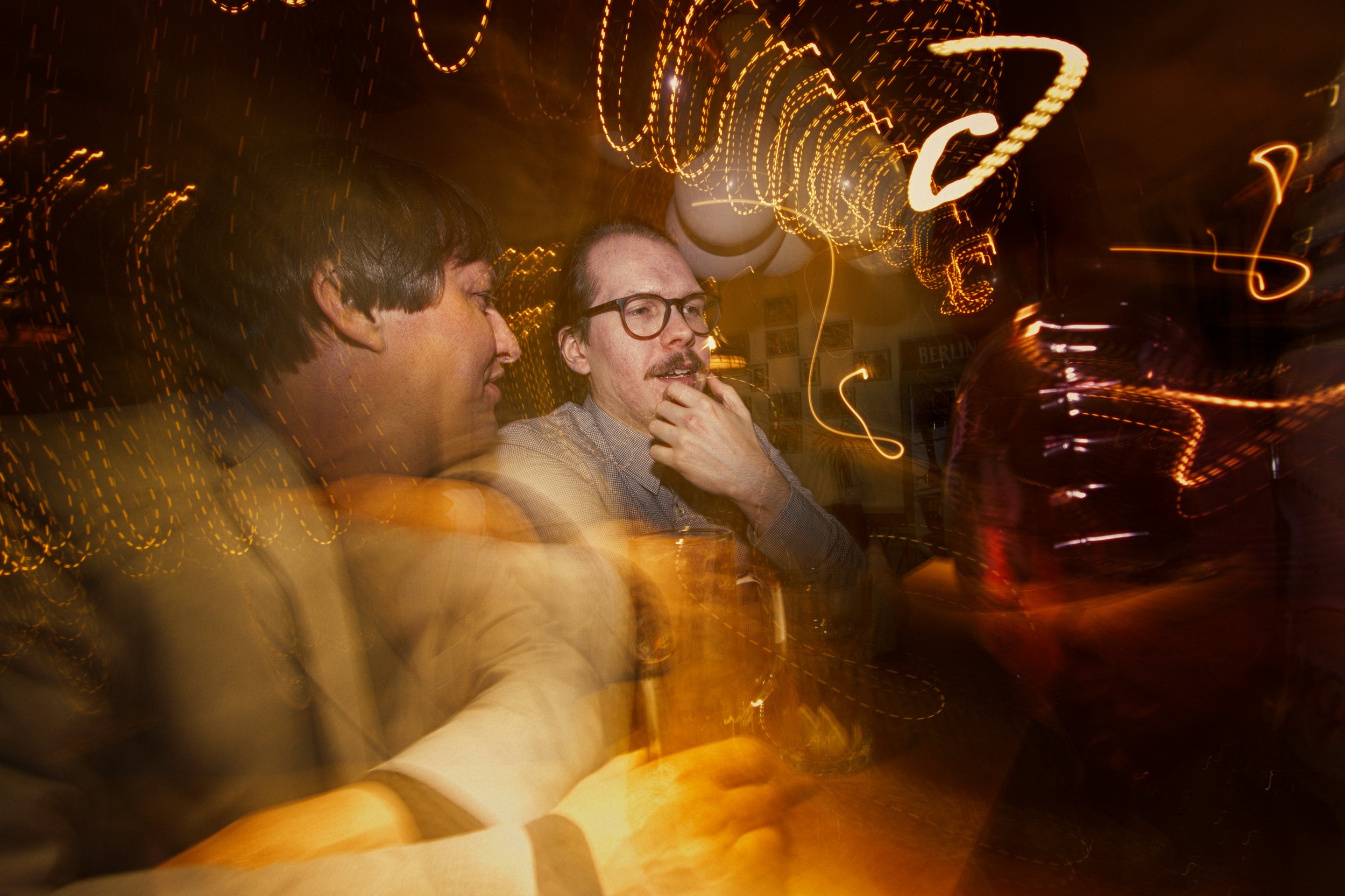Two men with glasses sitting at a table in a dimly lit venue with swirling orange lights and motion blur, possibly a bar or nightclub.