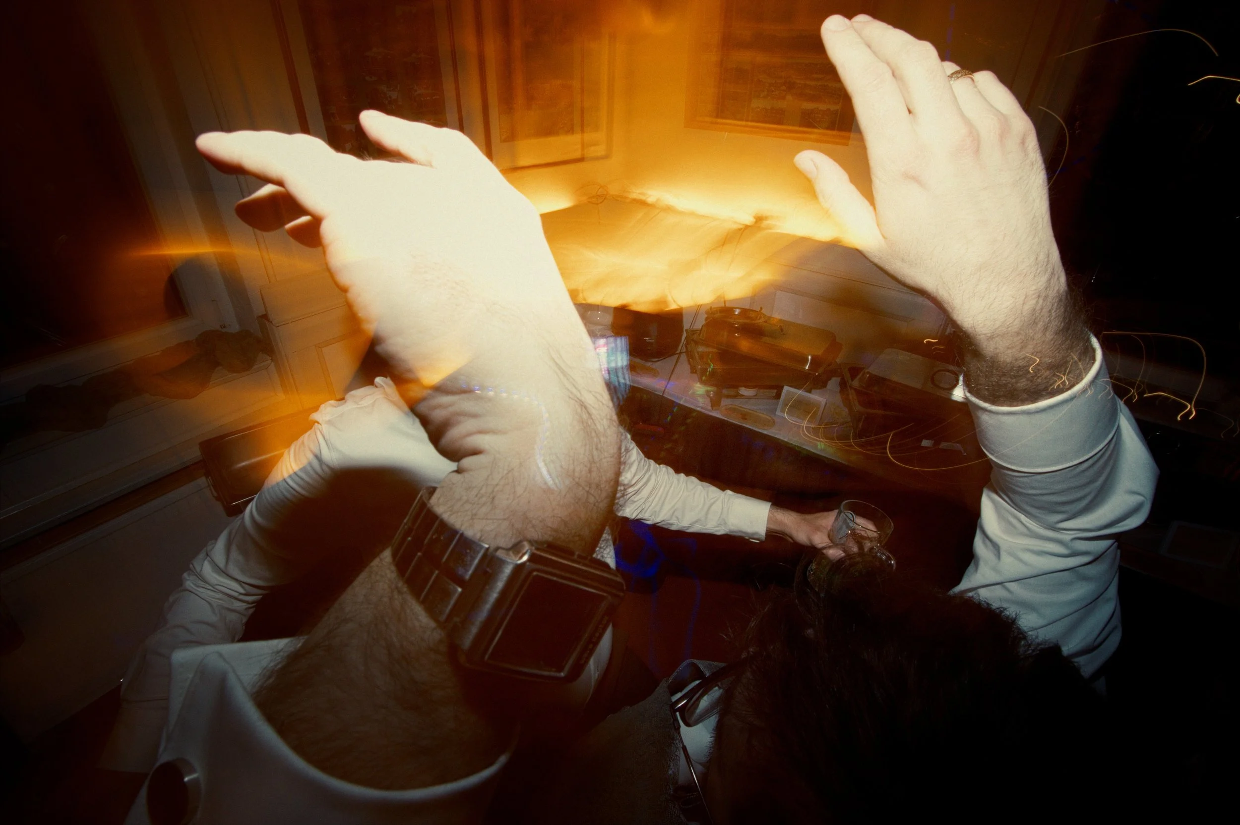 Hands raised in the air at a party or celebration, with a person holding a glass of drink sitting at a table in the background.