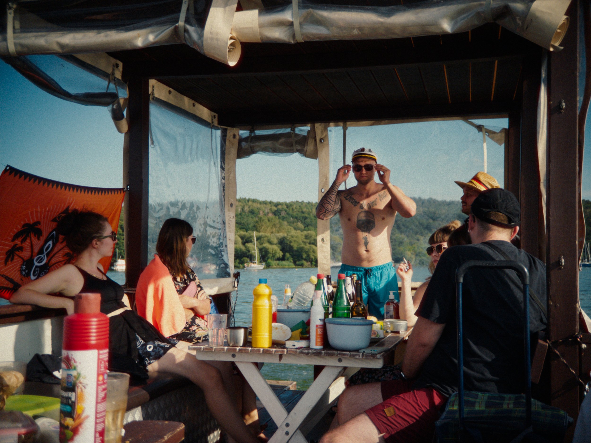 Group of friends on a boat enjoying a summer day. One man is standing shirtless, wearing sunglasses and a captain's hat, adjusting his sunglasses. The others are sitting around a table with drinks and snacks, some wearing hats and sunglasses, with a 