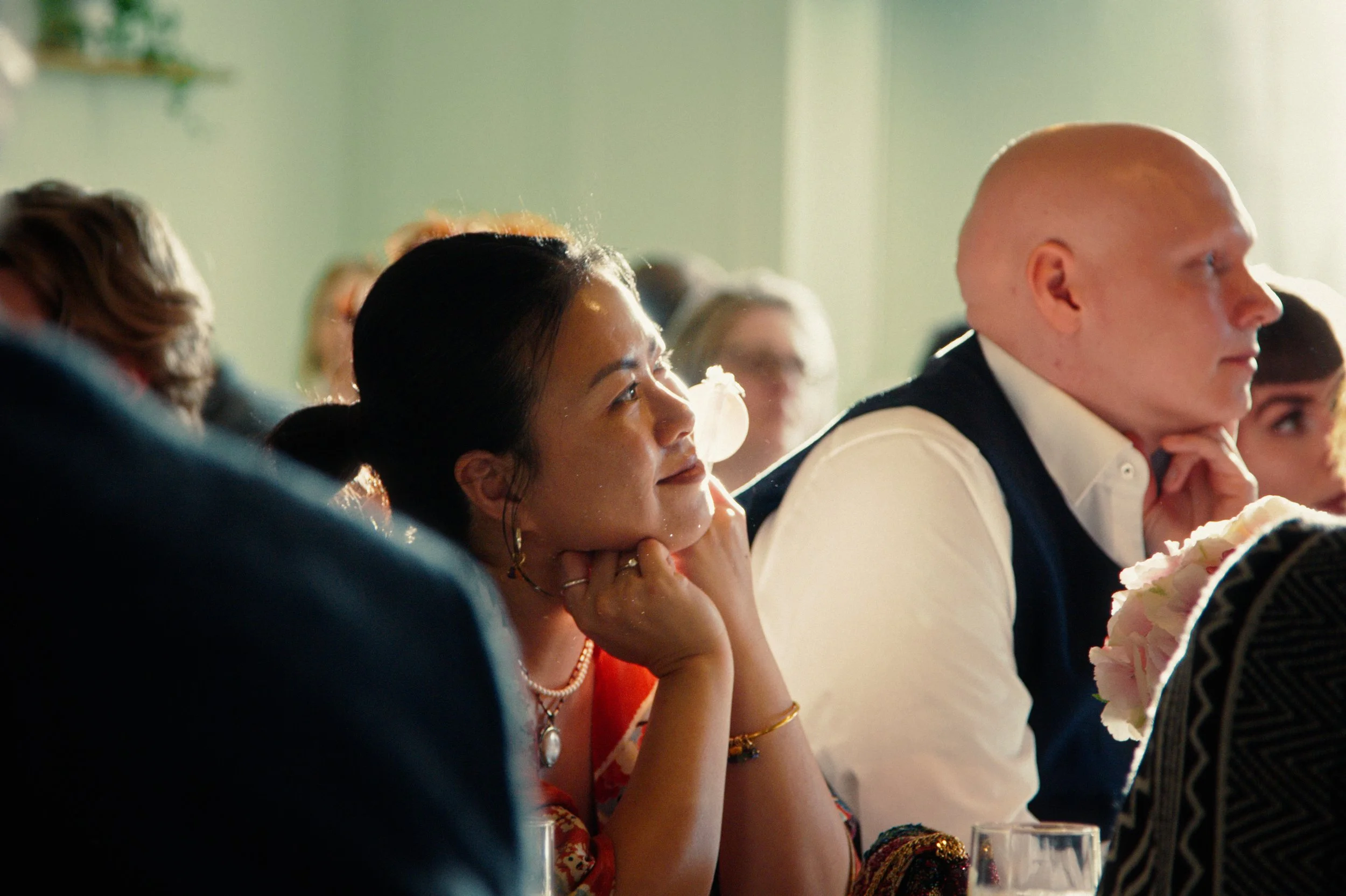 People participating in a formal event or gathering, sitting at a table, with some resting their chins on their hands, attentively listening.