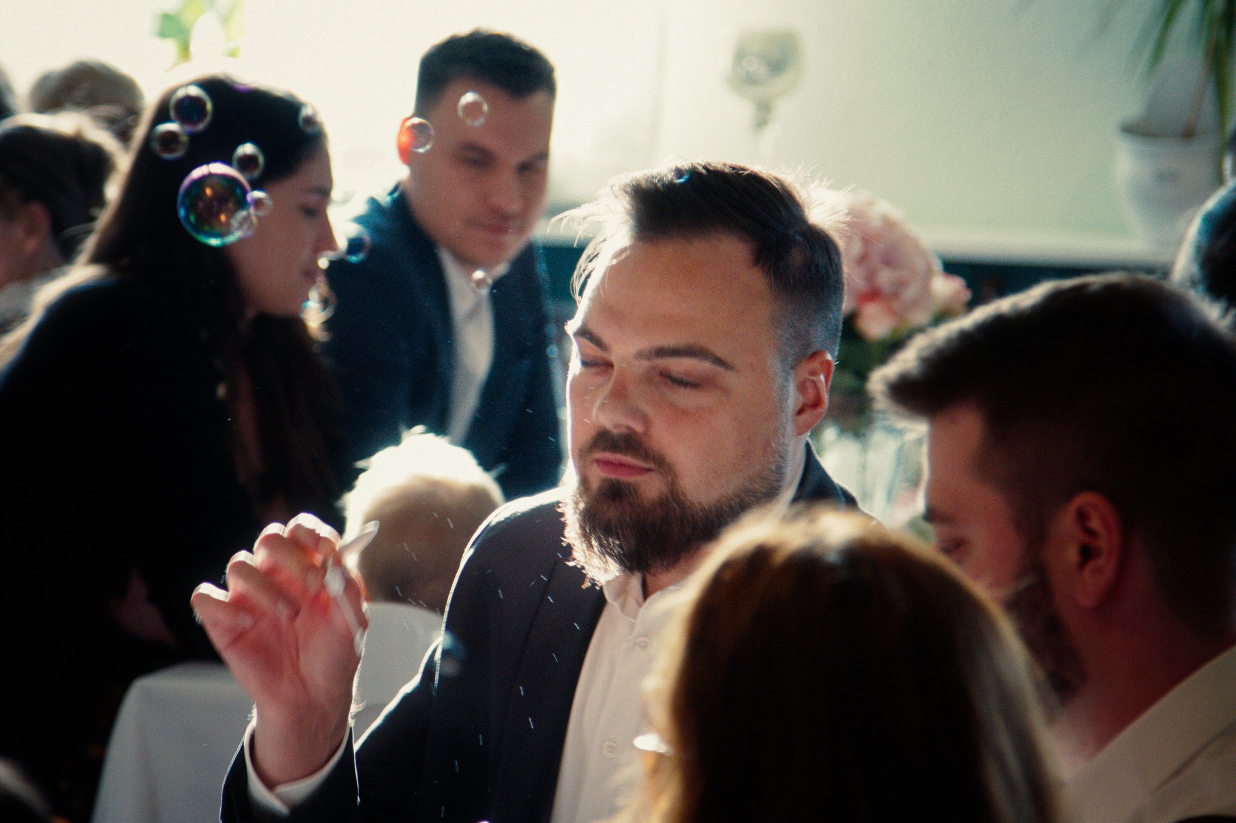 People socializing at a gathering or event, with sunlight streaming in, some blowing bubbles.