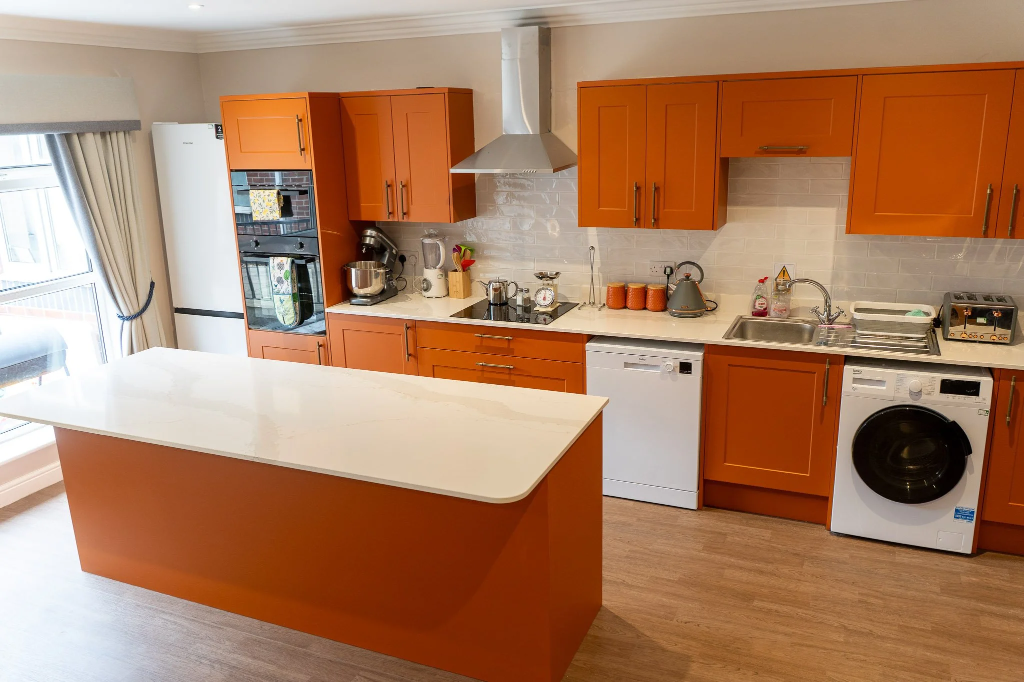 Kitchen with orange cabinets, white countertop, appliances including a built-in oven, microwave, dishwasher, washing machine, and a stove with a stainless steel range hood. Various kitchen items are on the counter, including a kettle, blender, and jars. There is a window with beige curtains on the left side.