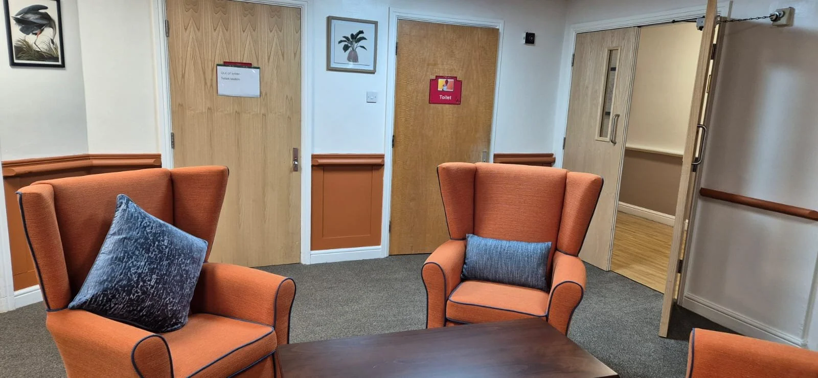 A cozy waiting room with three orange armchairs, each with a dark gray pillow, around a wooden coffee table. There are two wooden doors, one marked as the toilet, and walls decorated with framed pictures and a sign indicating the toilet is out of service.