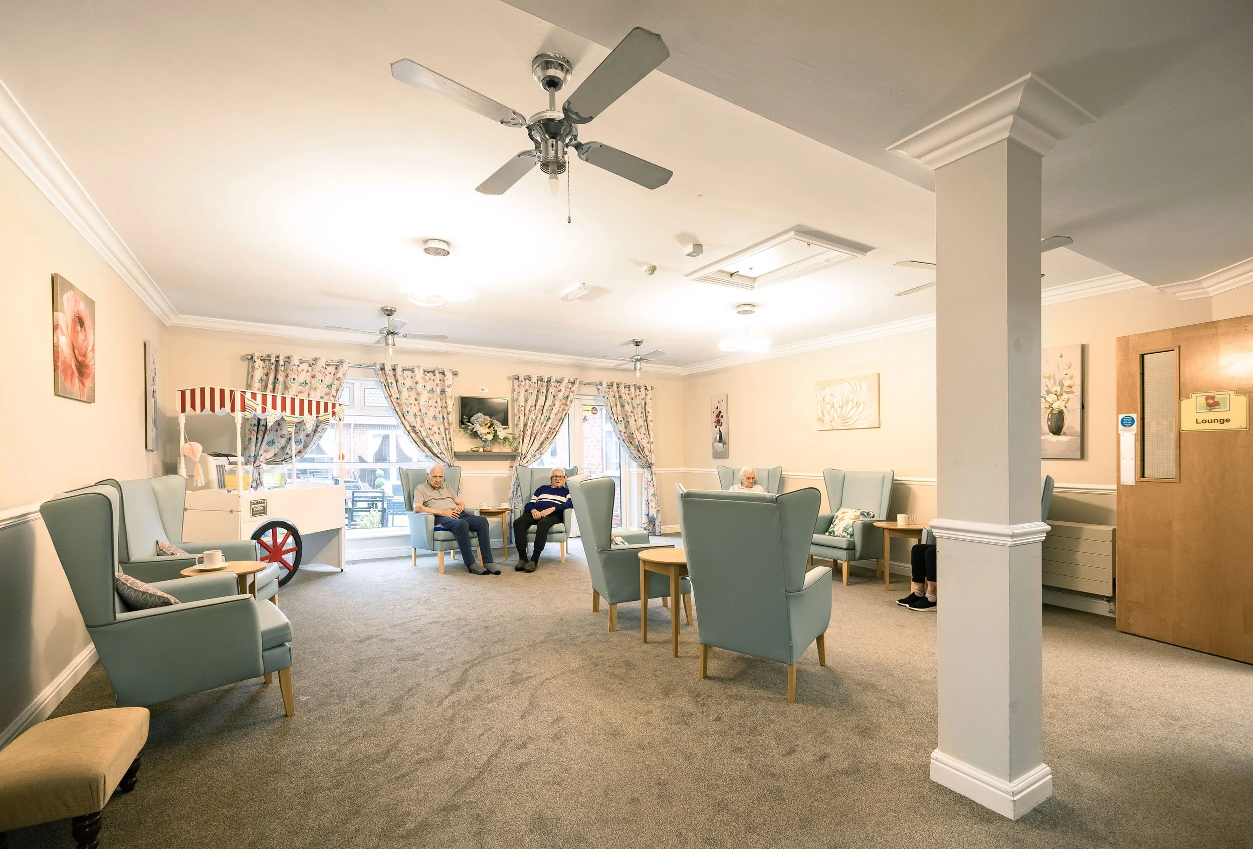 A spacious lounge area with light blue armchairs arranged in small groups and a few elderly people sitting on the chairs, decorated with floral curtains and paintings, with ceiling fans and natural light from windows.