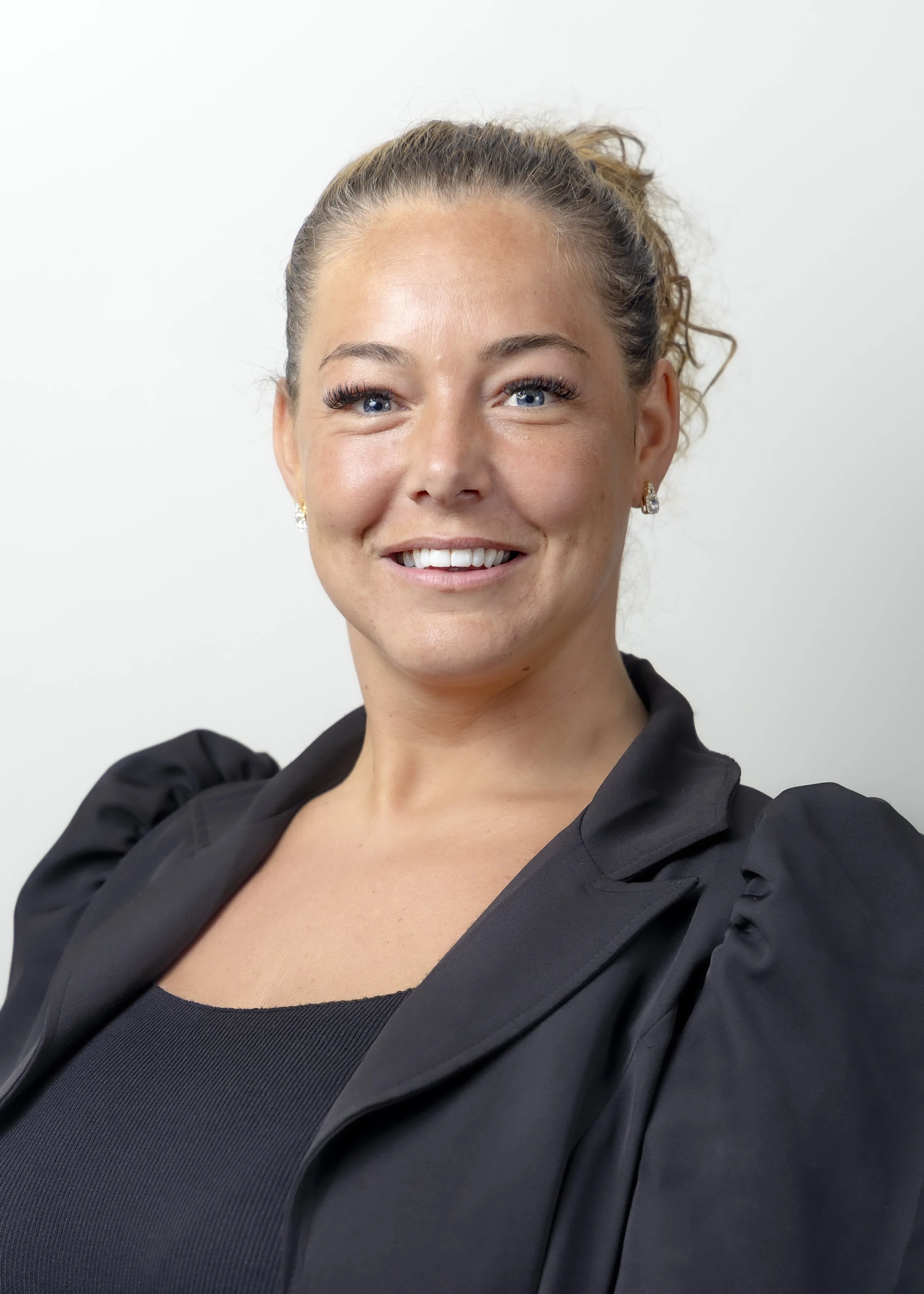 A woman smiling, with her hair in a bun, wearing earrings and a black top with puffed shoulders, standing against a plain white background.