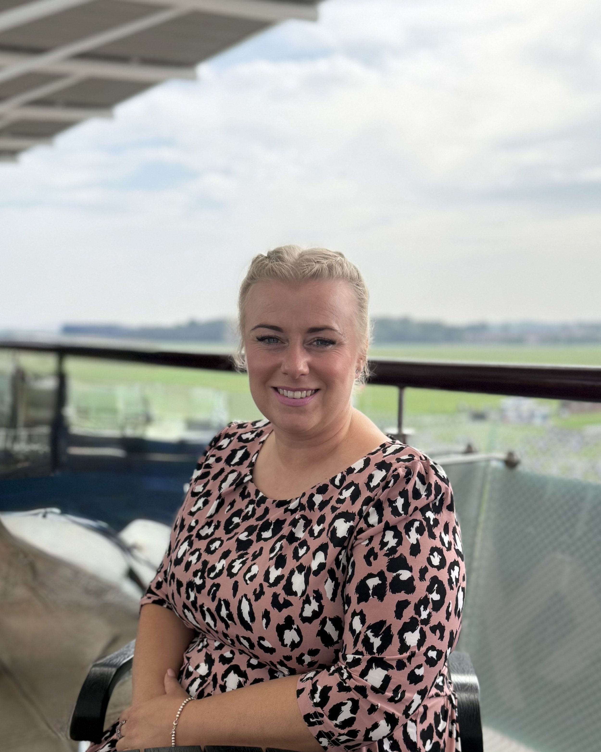 A woman with blonde hair smiling, wearing a leopard print top, sitting outdoors on a balcony or terrace with greenery and a cloudy sky in the background.