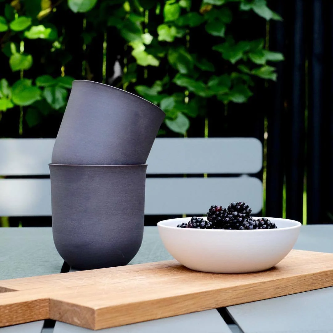 Handmade Scandinavian porcelain cup in black beside a white porcelain bowl filled with fresh blackberries on a wooden serving board outdoors.