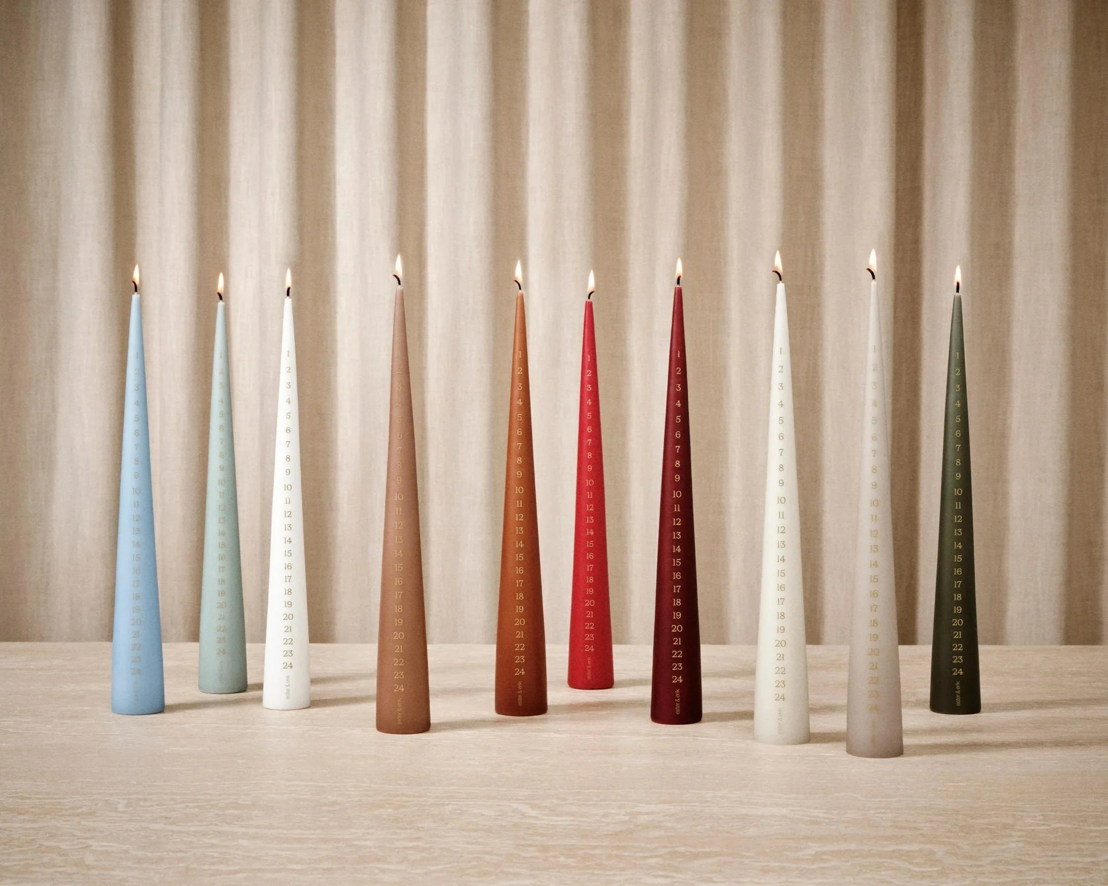 Ester & Erik cone calendar candles numbered 1–24 in assorted colours, lit and displayed together on a stone table.