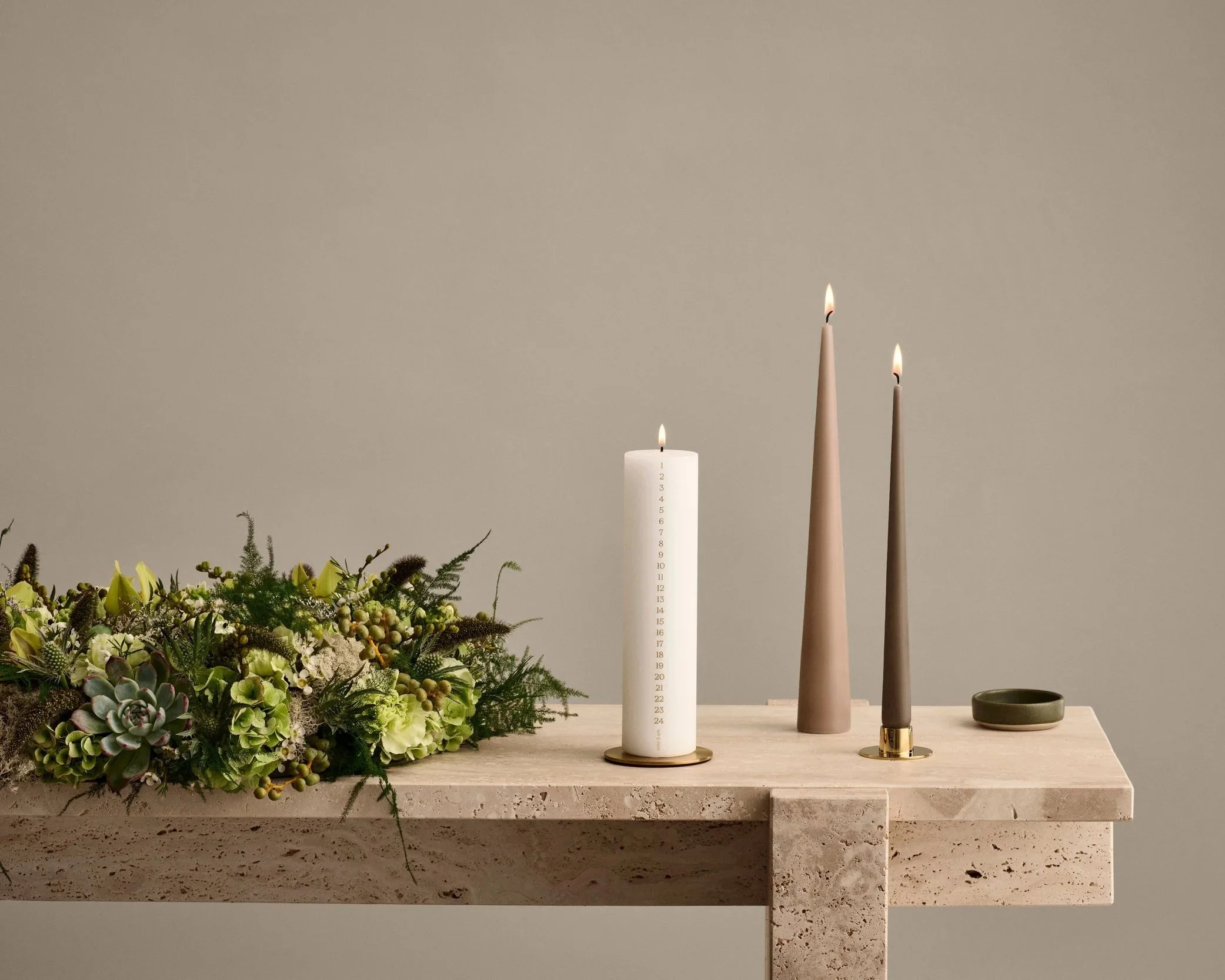 Ester & Erik white calendar candle numbered 1–24 with two lit cone candles and seasonal flowers on a stone table.