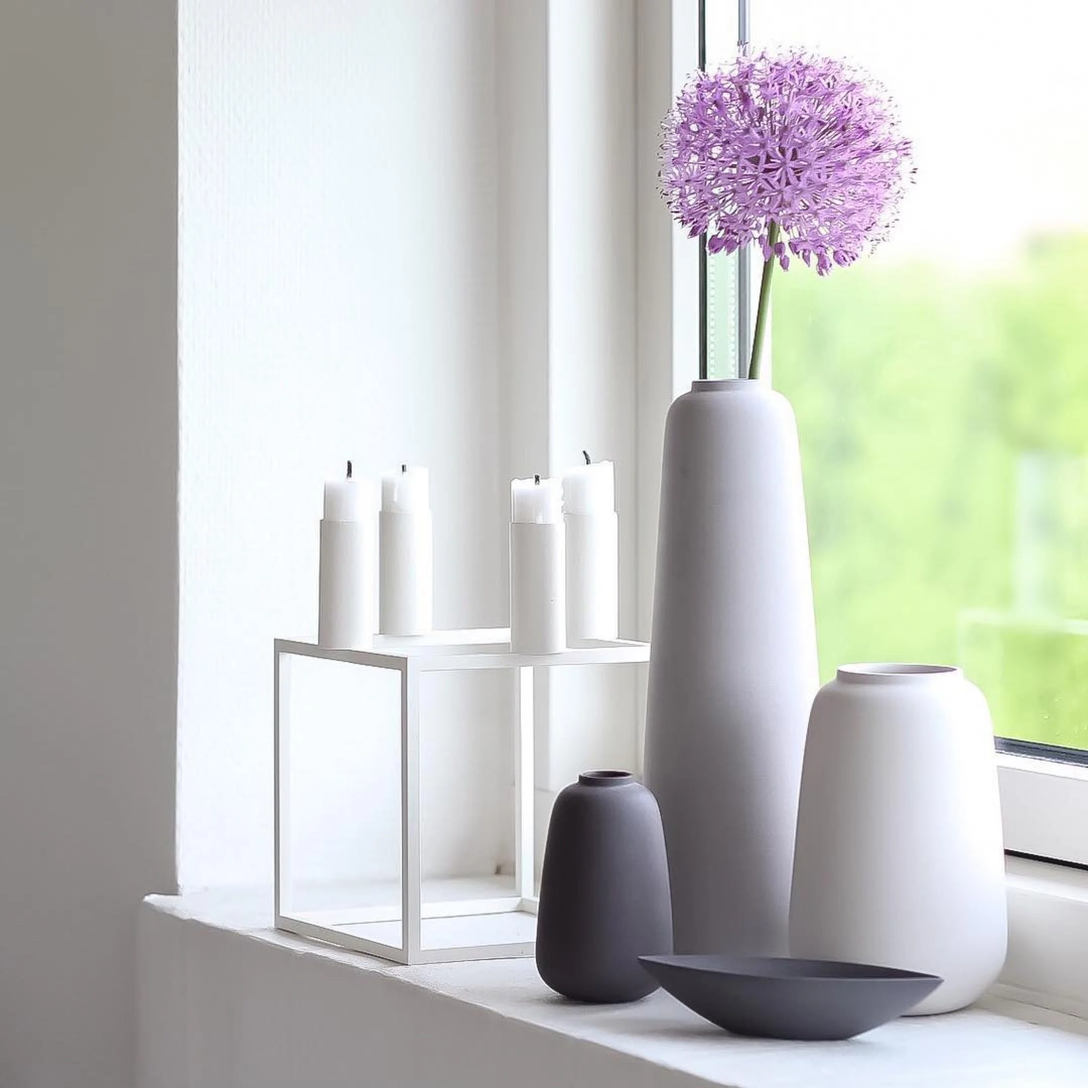 Handmade Scandinavian porcelain vases in white and black displayed on a windowsill with a purple allium flower, boat bowl, and white candle holders.