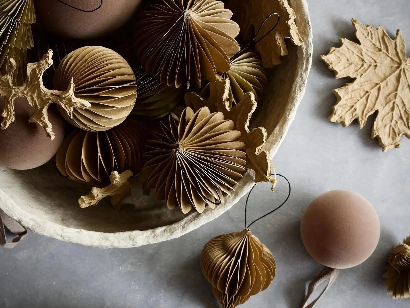 Folded paper Christmas ornaments in warm brown and natural tones, arranged in a rustic bowl with decorative leaves.