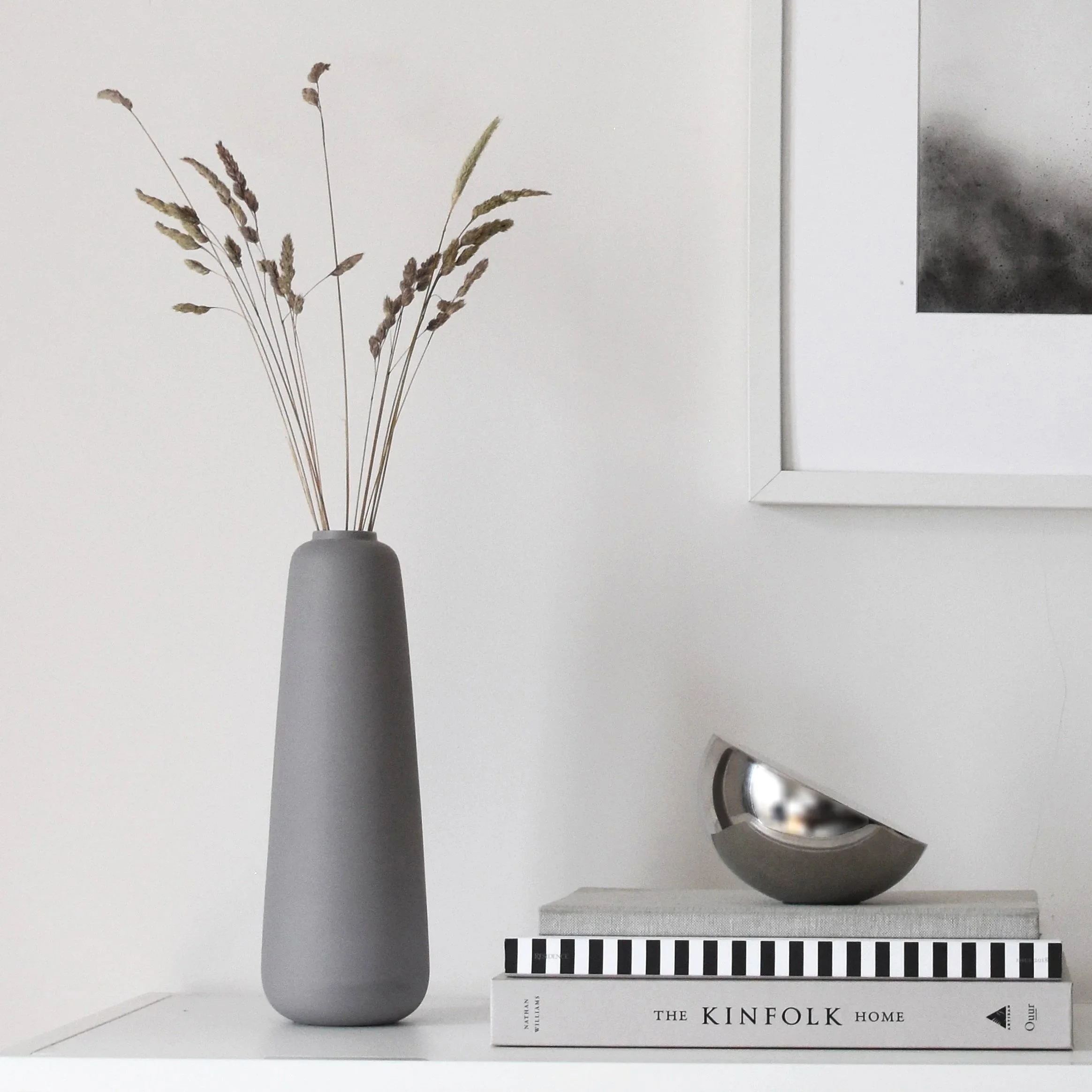 Tall grey ceramic vase with dried grasses, displayed alongside books and a sculptural metal object on a white shelf.