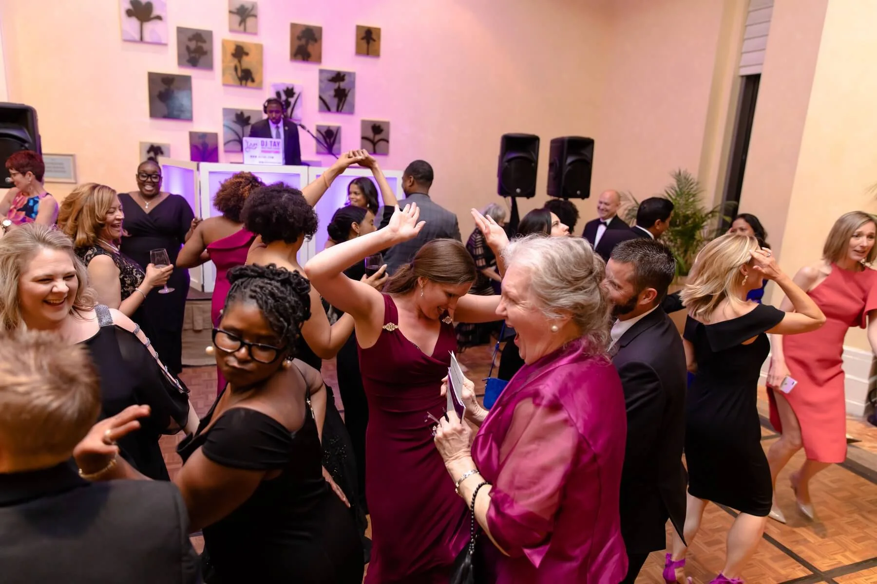 People dancing and socializing at a lively party with a DJ playing music in the background. The room has colorful lighting and floral artwork on the walls.