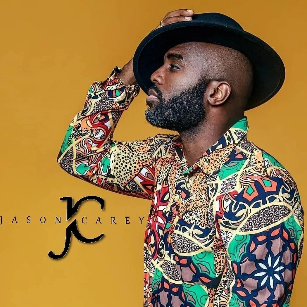 A man with a beard and shaved head wearing a colorful patterned shirt and a black hat, posing against a mustard yellow background.