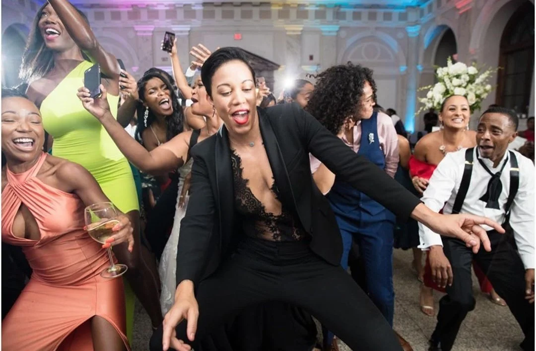 People dancing and having fun at a social event or party, with one woman holding a wine glass and others taking photos and smiling.