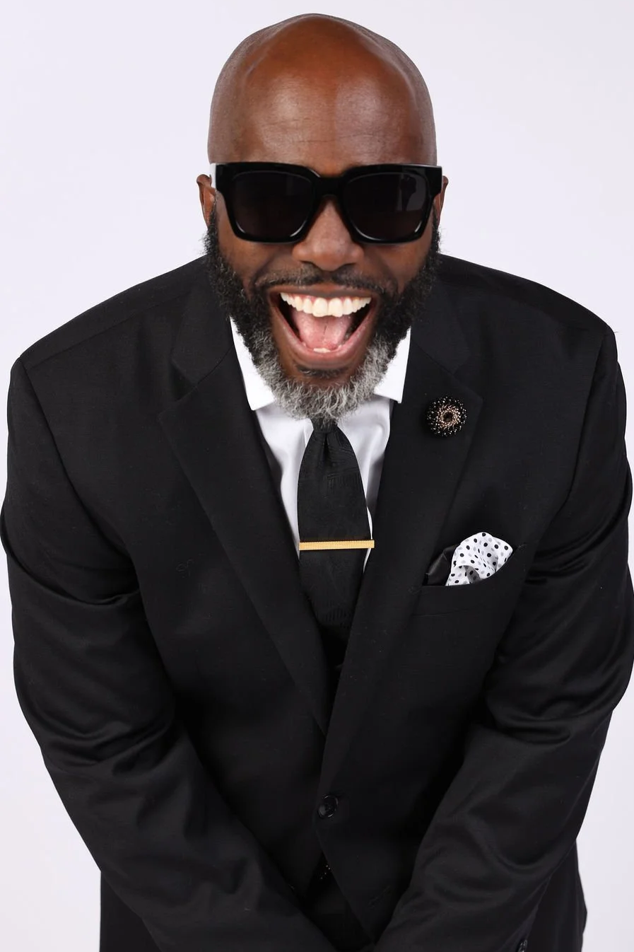 A bald African American man with a beard, wearing black sunglasses, a black suit, white shirt, black tie with a gold clip, and a white polka-dot pocket square, laughing joyfully with his mouth open and arms slightly bent.