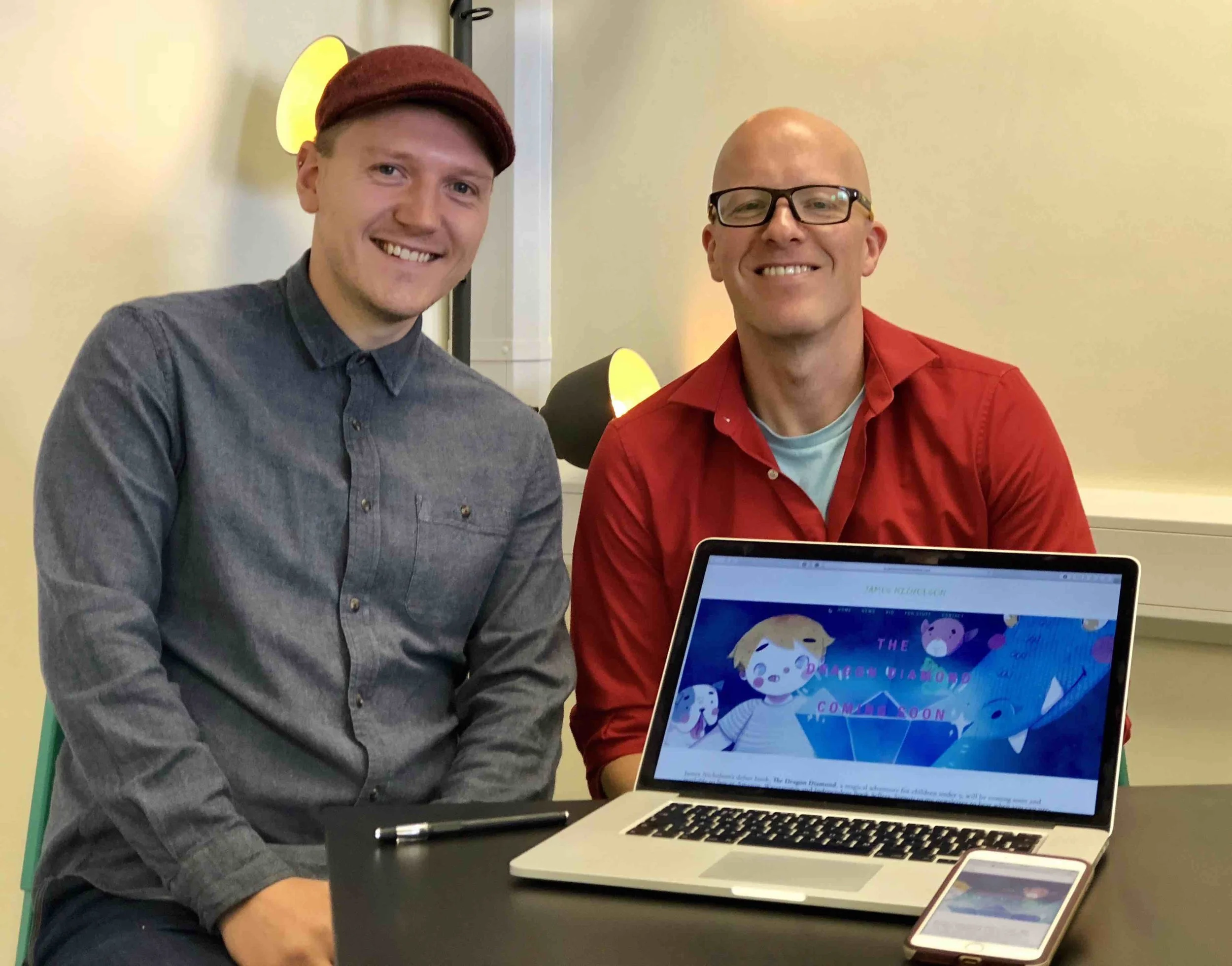 Young man with grey jumper smiling with red cap sitting next to another man wearing blue t shirt, red shirt, both smiling sitting around black table in office with website and phone of James Nicholson.com