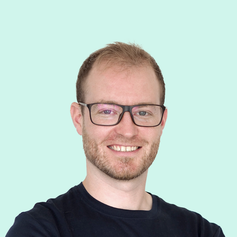 Jonny, a white male with short hair and beaming smile, wearing black rectangular frame glasses.