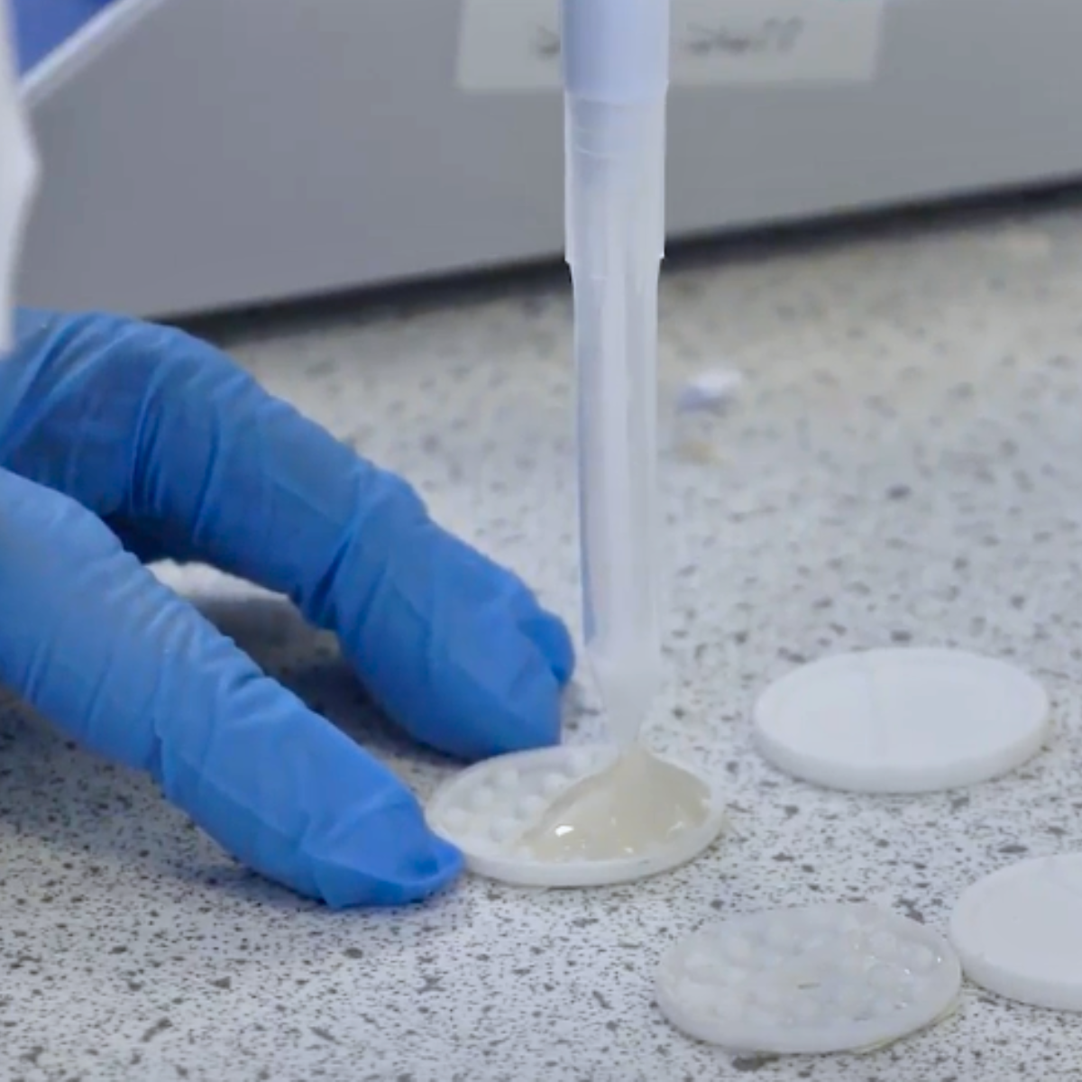 A close-up of hands in blue gloves pipetting a gel into a small disc tray.