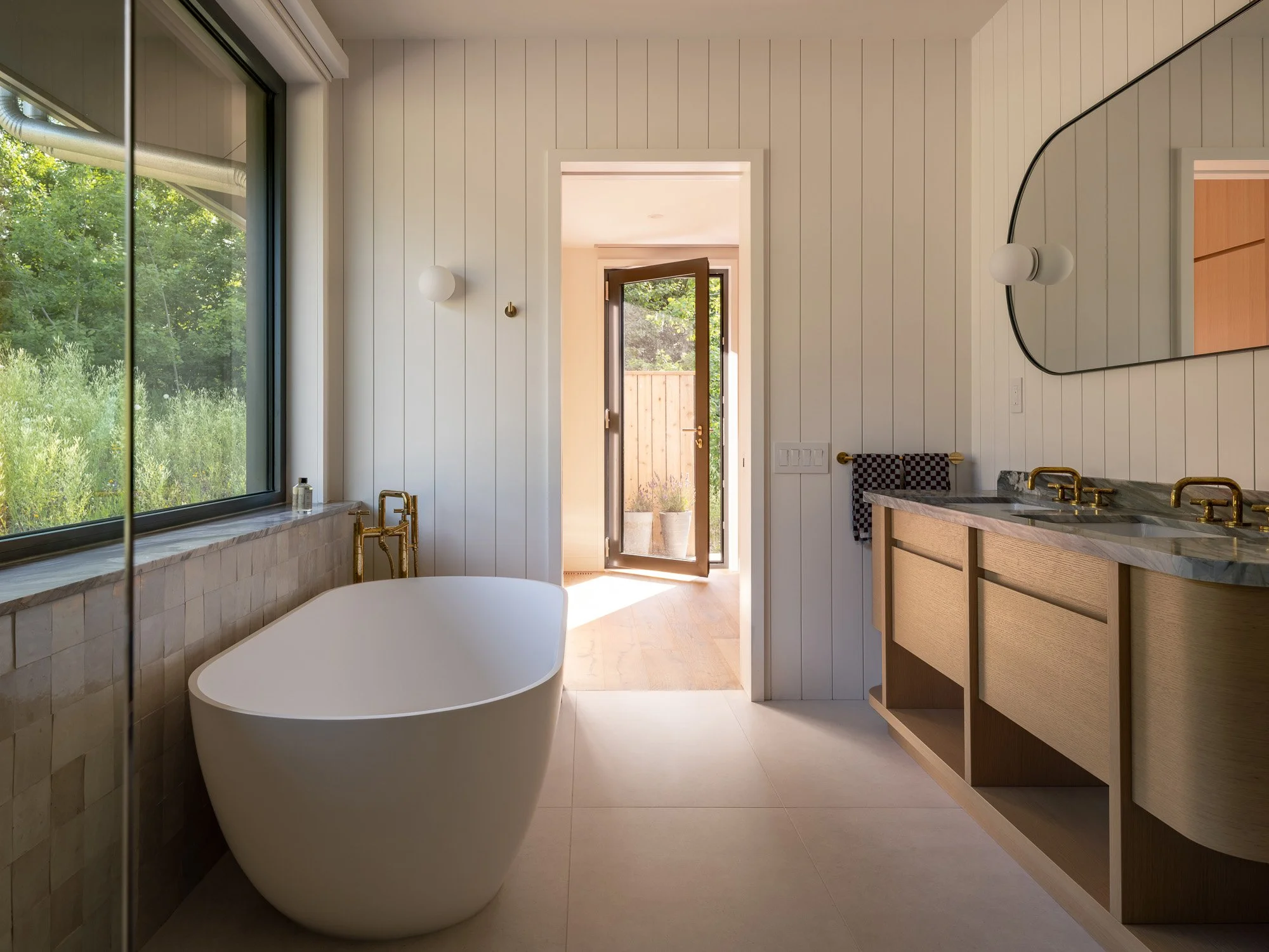 Luxurious primary bathroom suite featuring a freestanding soaking tub, organic wall tile, custom wood millwork, and a large window.