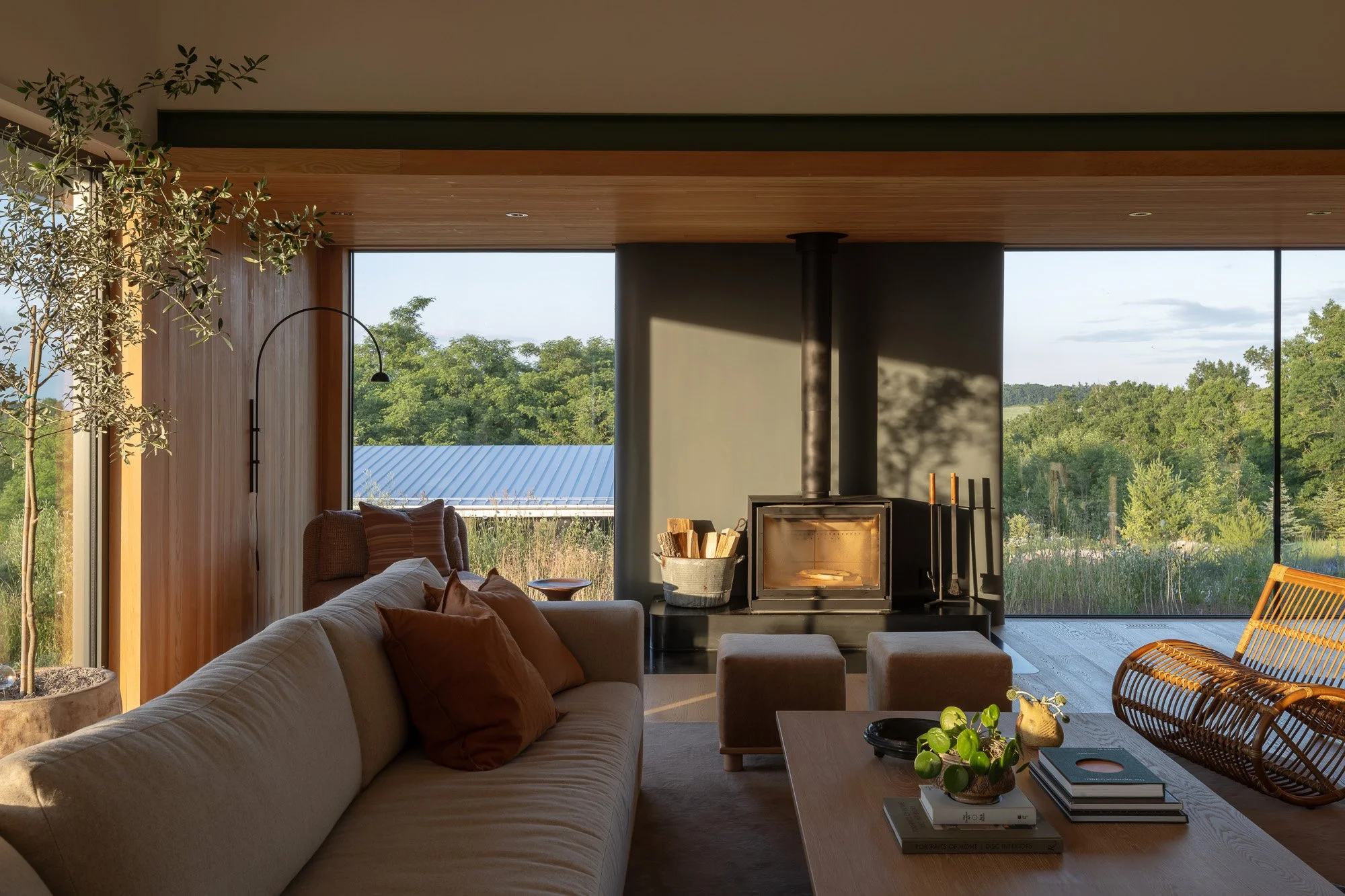 Warm modernist living room interior with a wood-burning stove, cozy family furniture, and large floor-to-ceiling windows. Minimalist open living area with sliding glass doors that drop flush into the floor to connect the interior with the surrounding