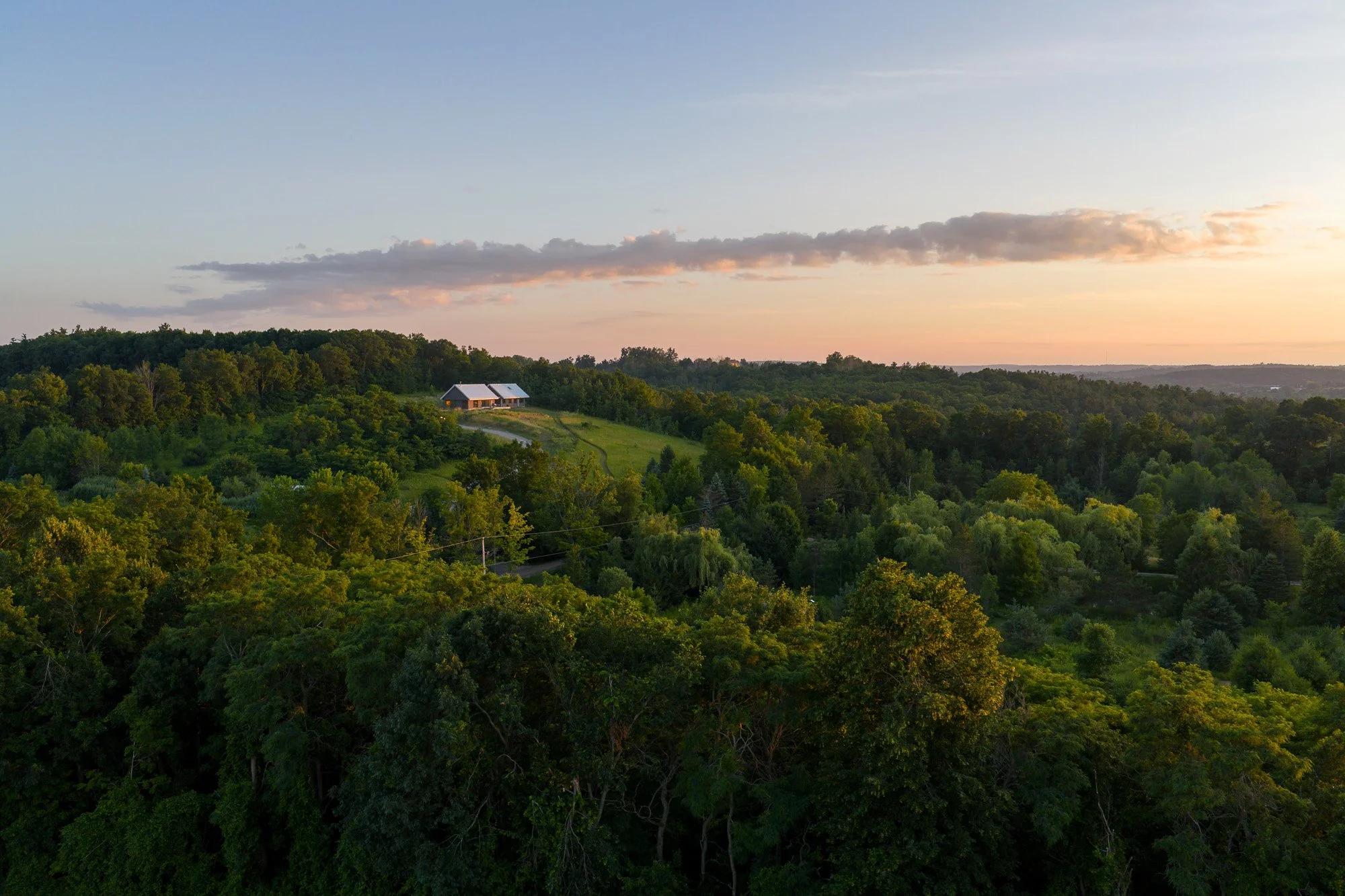 Modern rural home design by Wayback Architects featuring a unique curved gable roof overlooking the rolling hills of Northumberland County, Ontario during sunset.