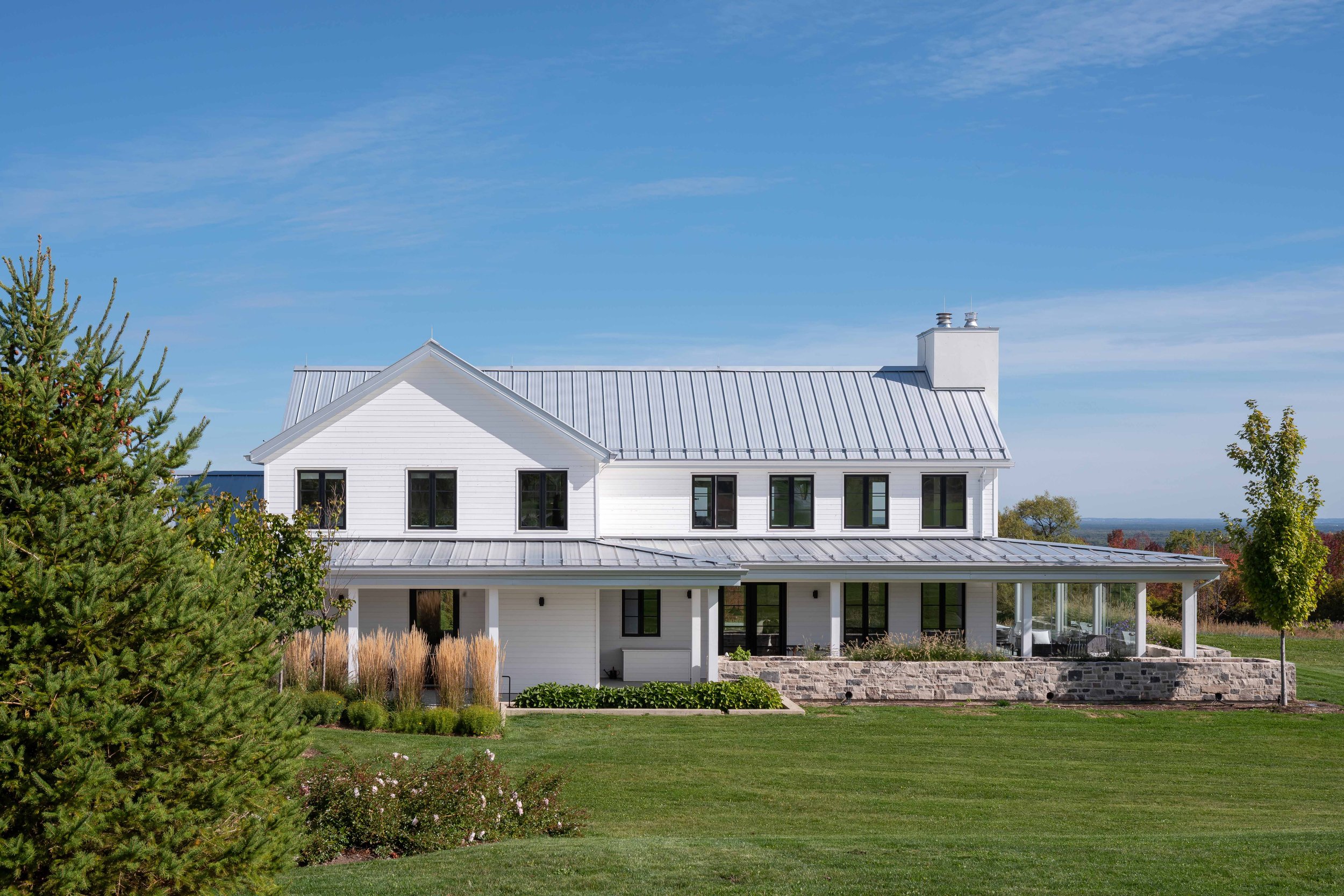 Modern farmhouse exterior showcasing an expansive wrap-around porch living space anchored by a natural stone planter base.