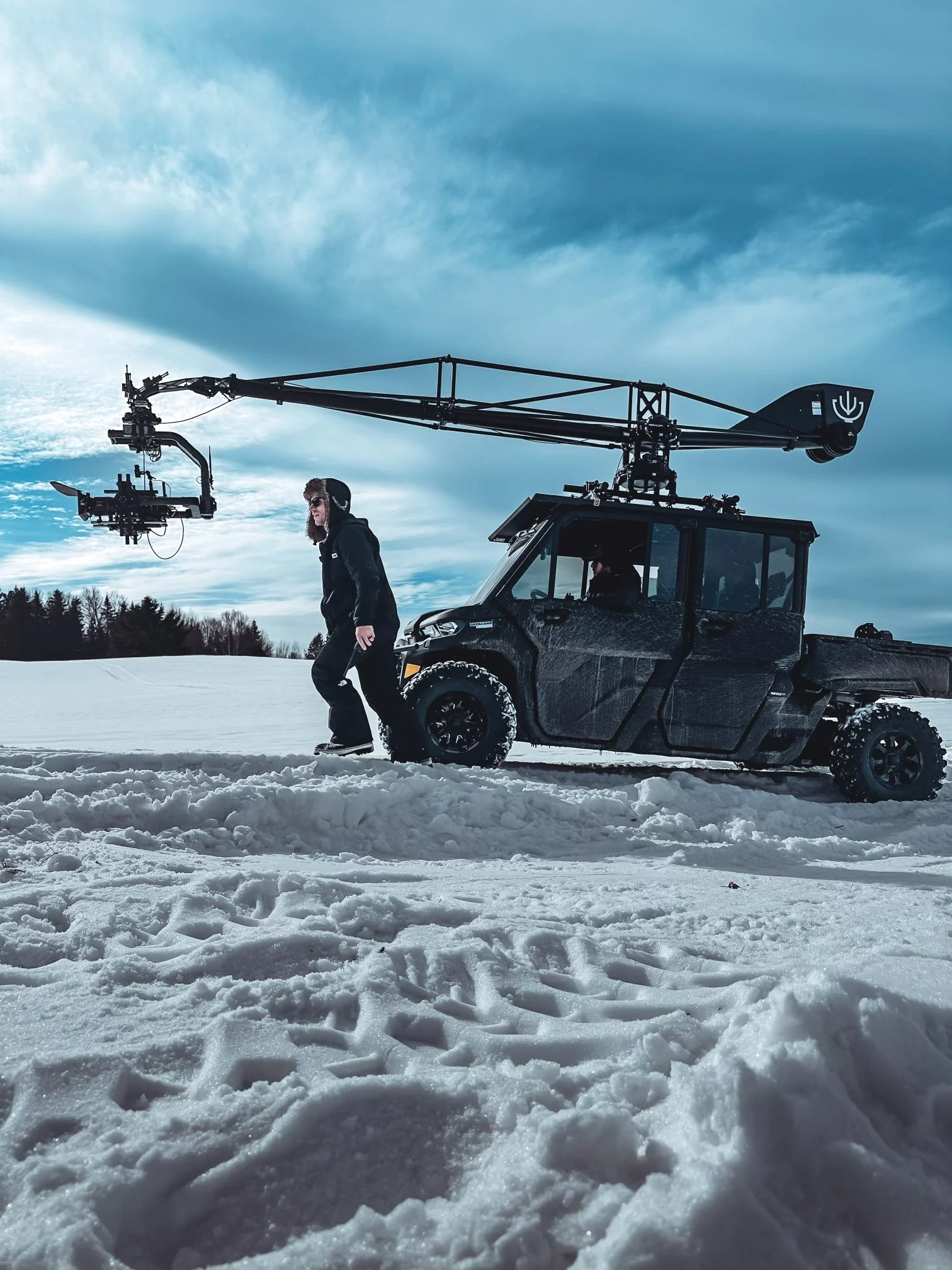 A crew member walks beside a camera-rigged utility vehicle in a snowy field.