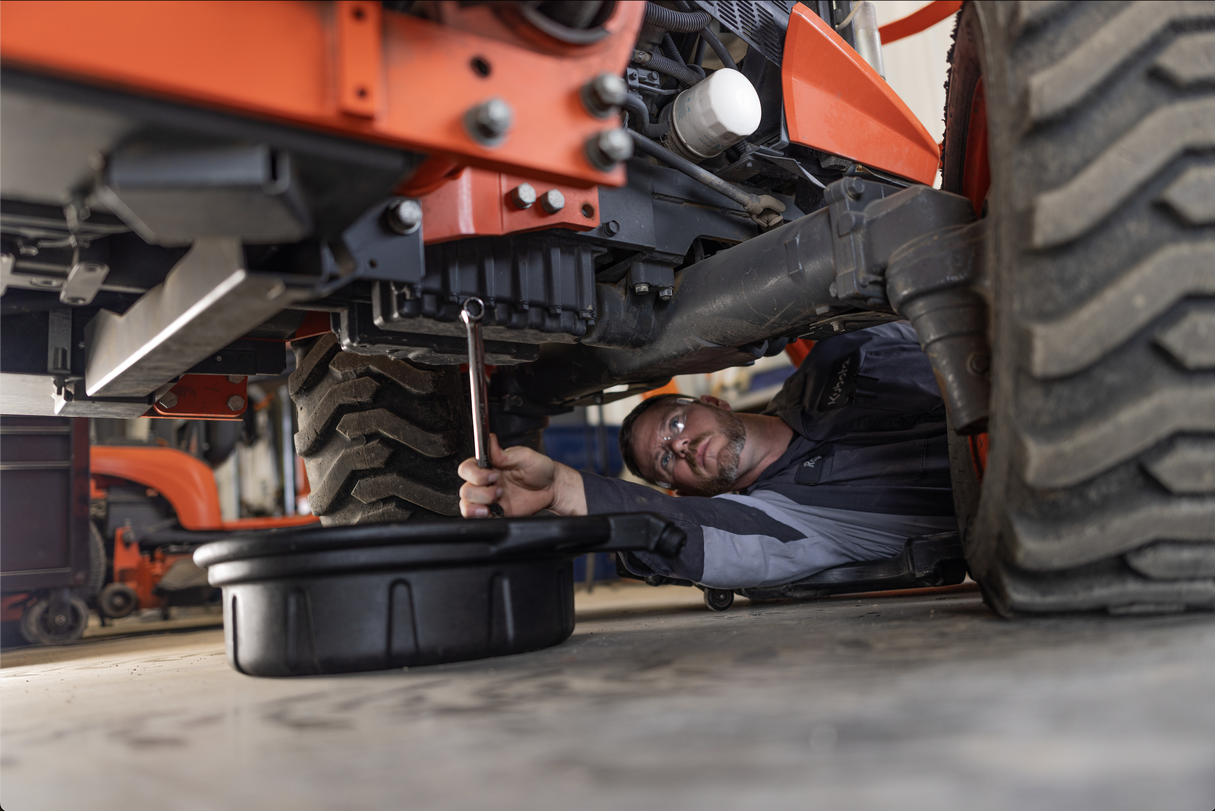 A mechanic works beneath orange farm equipment during agriculture video production.