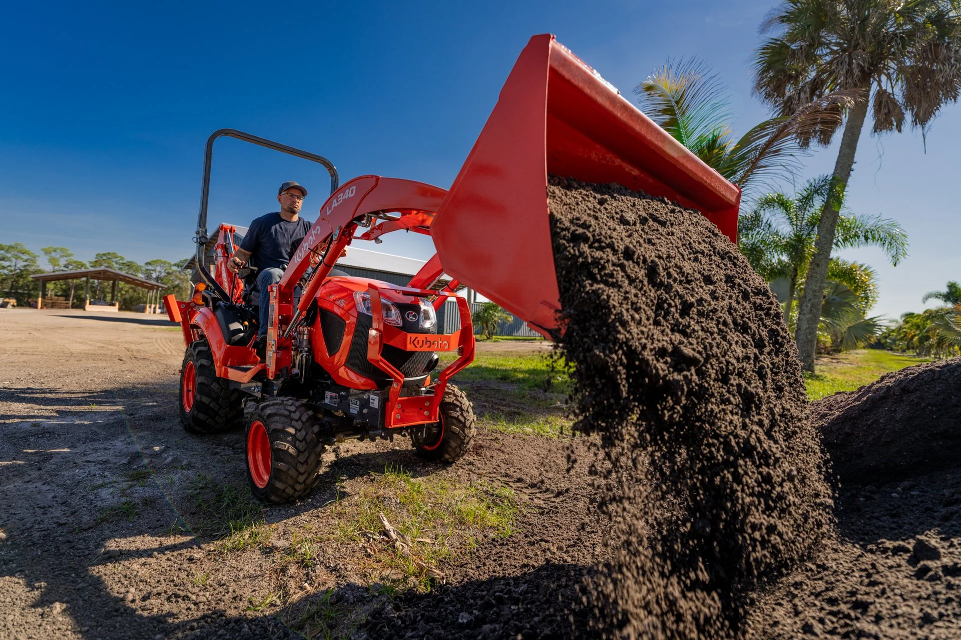 A front loader tips dark soil onto a garden bed in a sunny equipment video production scene.